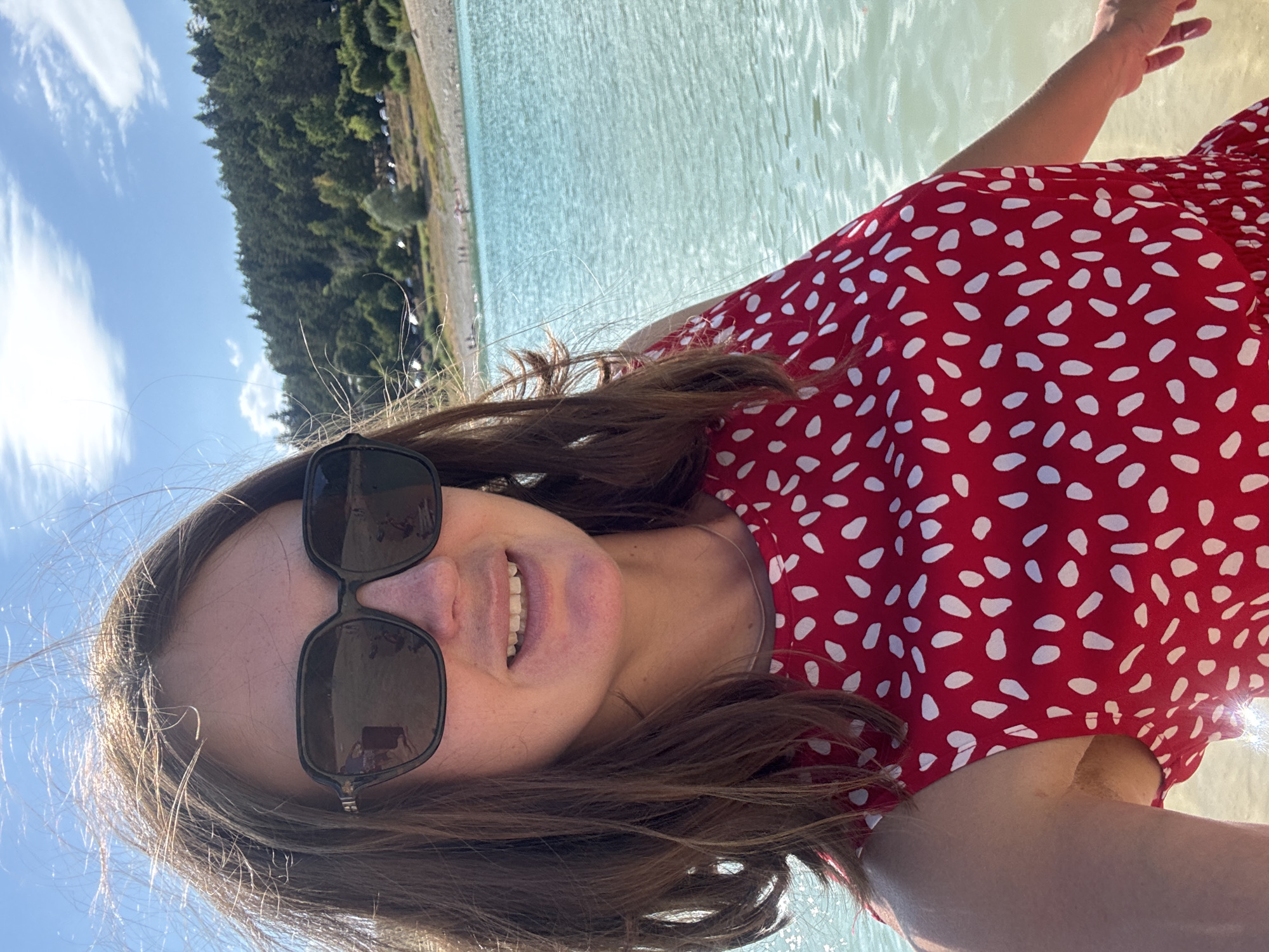 A woman, wearing a red dress with white polka dots and large black sunglasses, is captured in a selfie outdoors. She stands near a body of turquoise water, possibly a lake, with a shoreline populated by a few individuals and a parking area visible in the background. Beyond the shoreline, a dense forest covers rolling hills under a bright blue sky with scattered clouds. The scene suggests a recreational area, likely the Lac de Vouglans, located near the commune of Maisod in the Jura department of France. The lighting indicates a sunny day, with some wind causing the woman's hair to blow across her face.