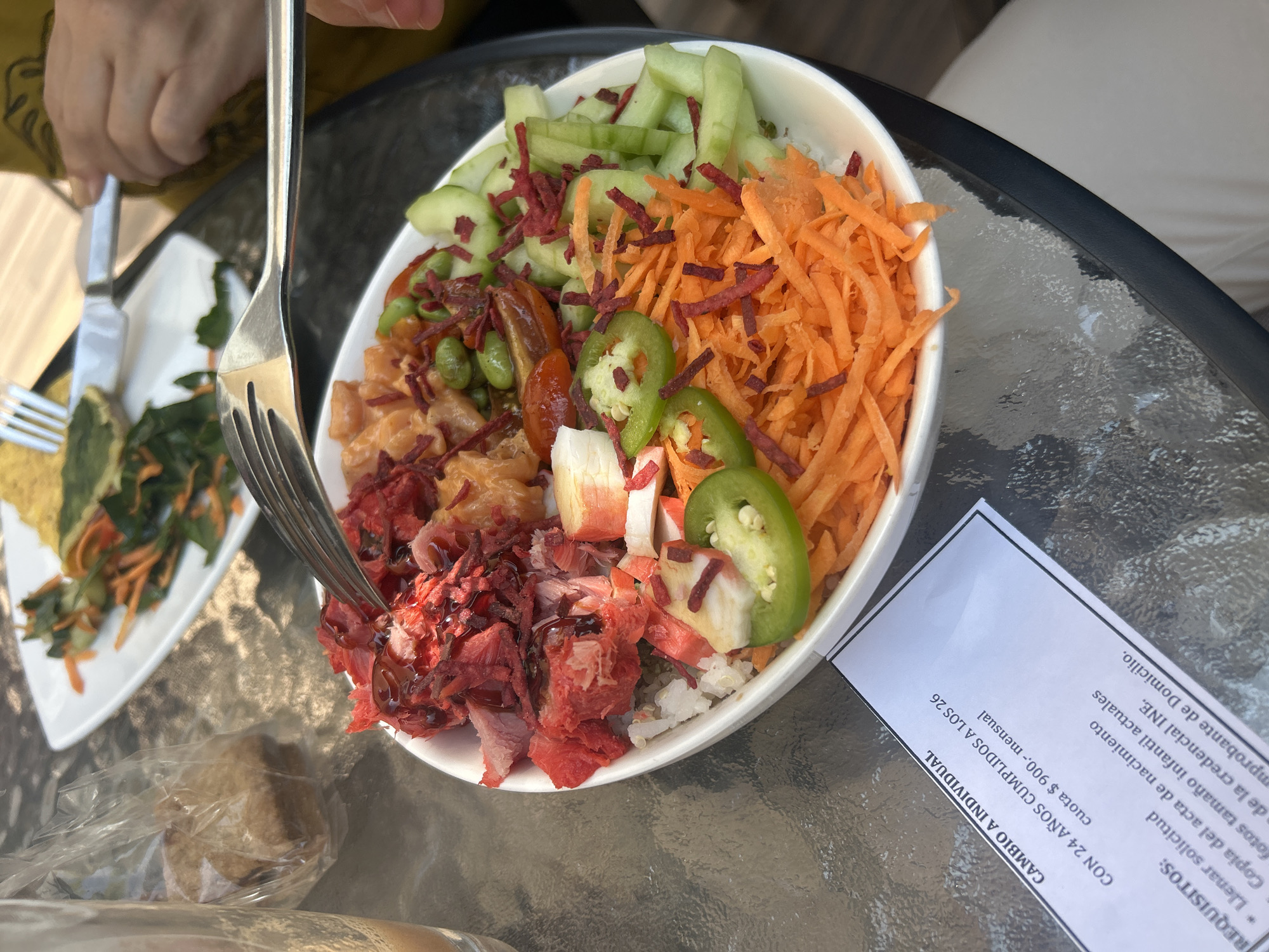 The image shows a partially eaten poke bowl on a glass table. The bowl contains rice, red tuna with a dark sauce, salmon, edamame, cucumber slices, shredded carrots, crab sticks, and jalapeno slices. A fork is piercing the tuna in the bowl. To the left, there is a second plate with what appears to be a salad with similar ingredients. Next to the poke bowl, there is a piece of bread in a plastic bag and a sheet of paper with text in Spanish, indicating a change to individual fees. The text also mentions the age range of 24-26 years. The location of San Bernardino Tlaxcalancingo, Mexico, cannot be determined from the image.