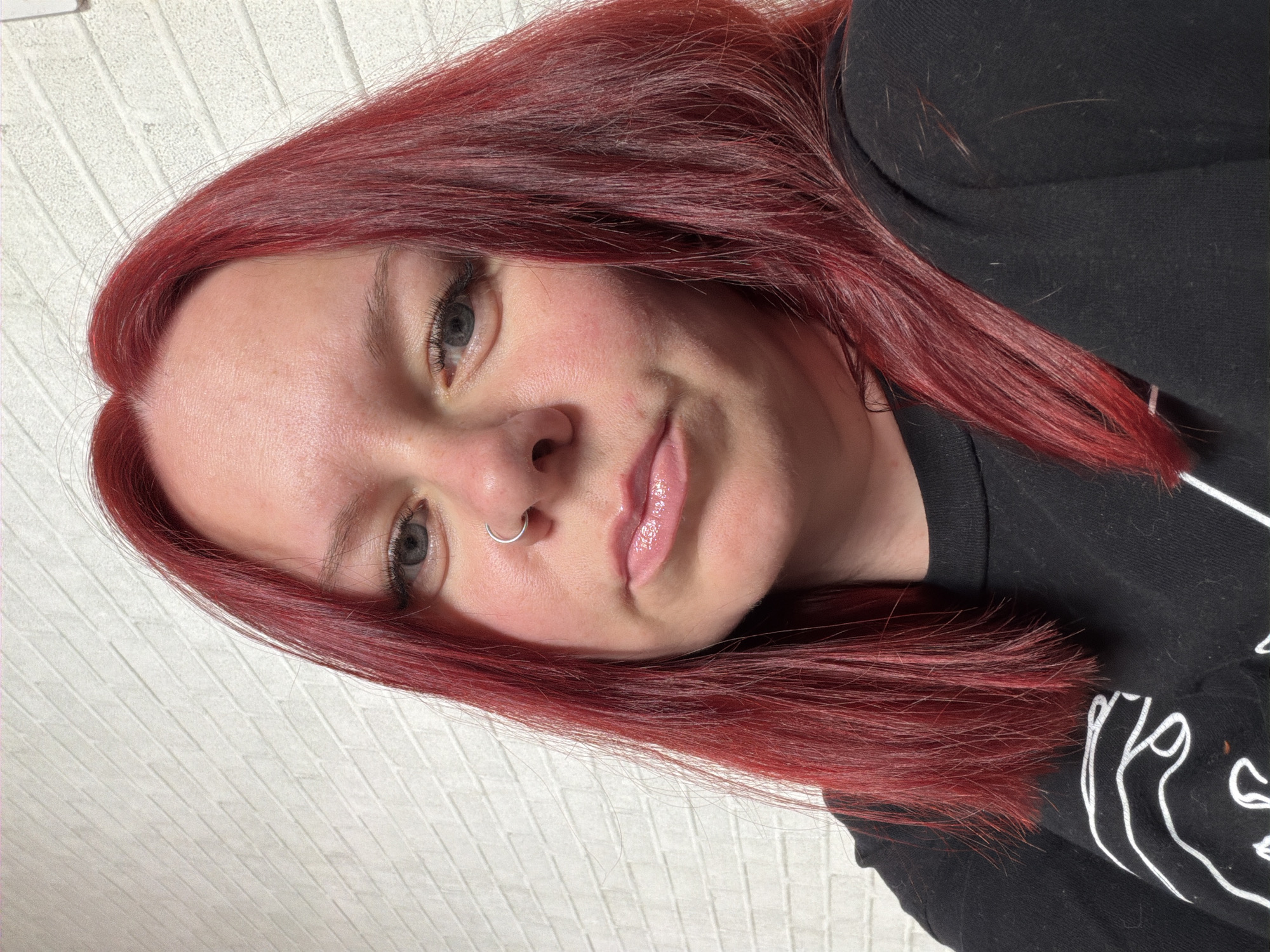 A female subject is depicted in a close-up portrait, facing forward and making direct eye contact with the camera. She has long, straight, deep red hair, parted slightly to her left, with some darker roots visible. Her complexion is fair, exhibiting some redness on her cheeks and forehead. She has blue-grey eyes and a silver-colored hoop piercing in her left nostril. Her lips are glossy and slightly parted, with the corners subtly downturned. She is wearing a black long-sleeved garment, possibly a sweatshirt, with a partial white, abstract graphic design visible on the left side of her chest. The background is a uniformly lit, light-colored wall featuring a textured pattern resembling white rectangular bricks arranged in a running bond configuration. The specific location within the city of Chaikovsky, Russia, is not discernible from the visual information in the image.