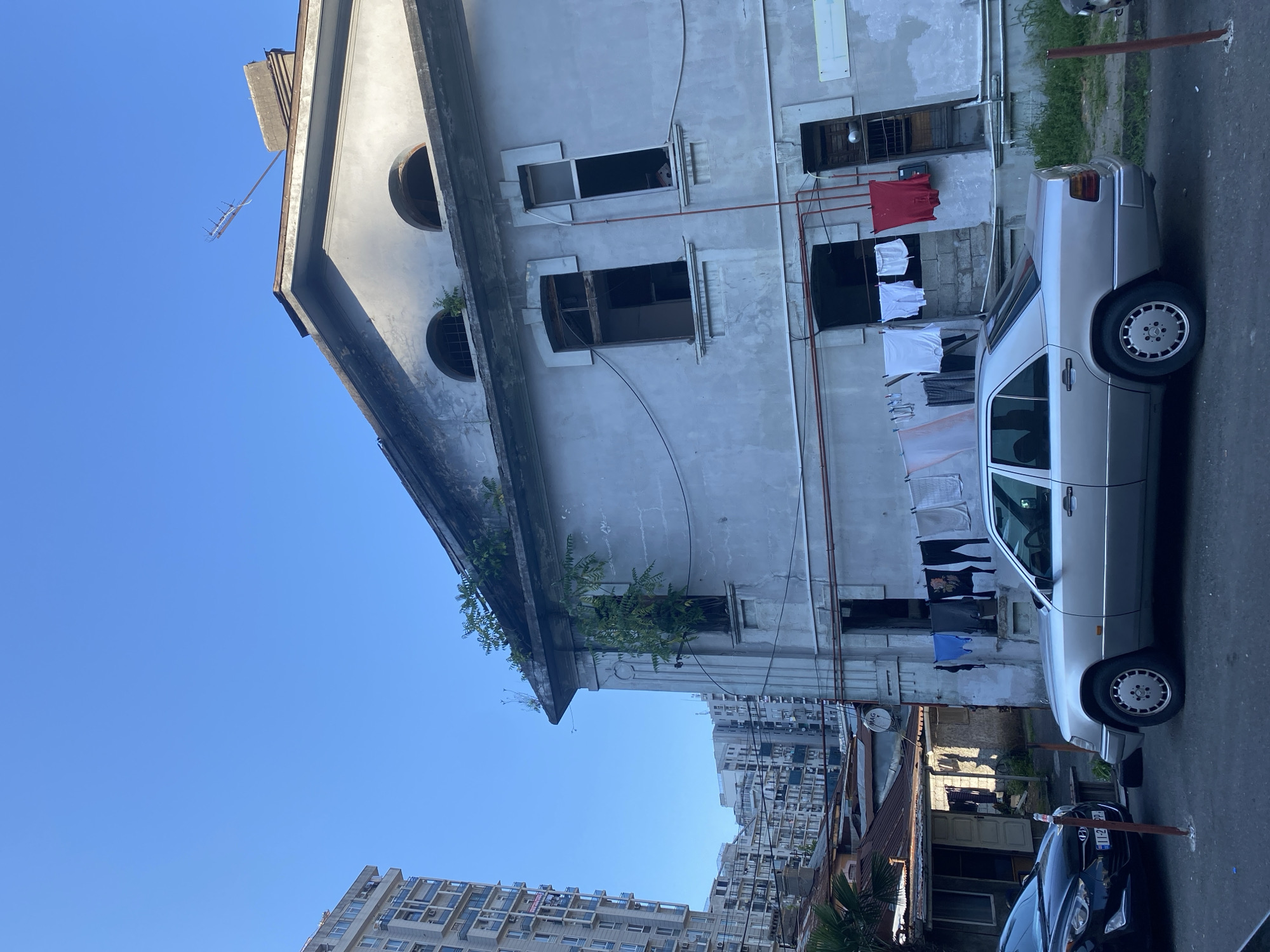 In Batumi, Georgia, a silver Mercedes-Benz W124 sedan is parked on a paved street in front of an old, weathered concrete building with a grey facade. The building has several arched windows and a sloping roof with an antenna on top. Laundry is hanging on a clothesline stretched across the building's facade. In the background, modern high-rise apartment buildings are visible, contrasting with the older architecture. The sky is clear and blue.