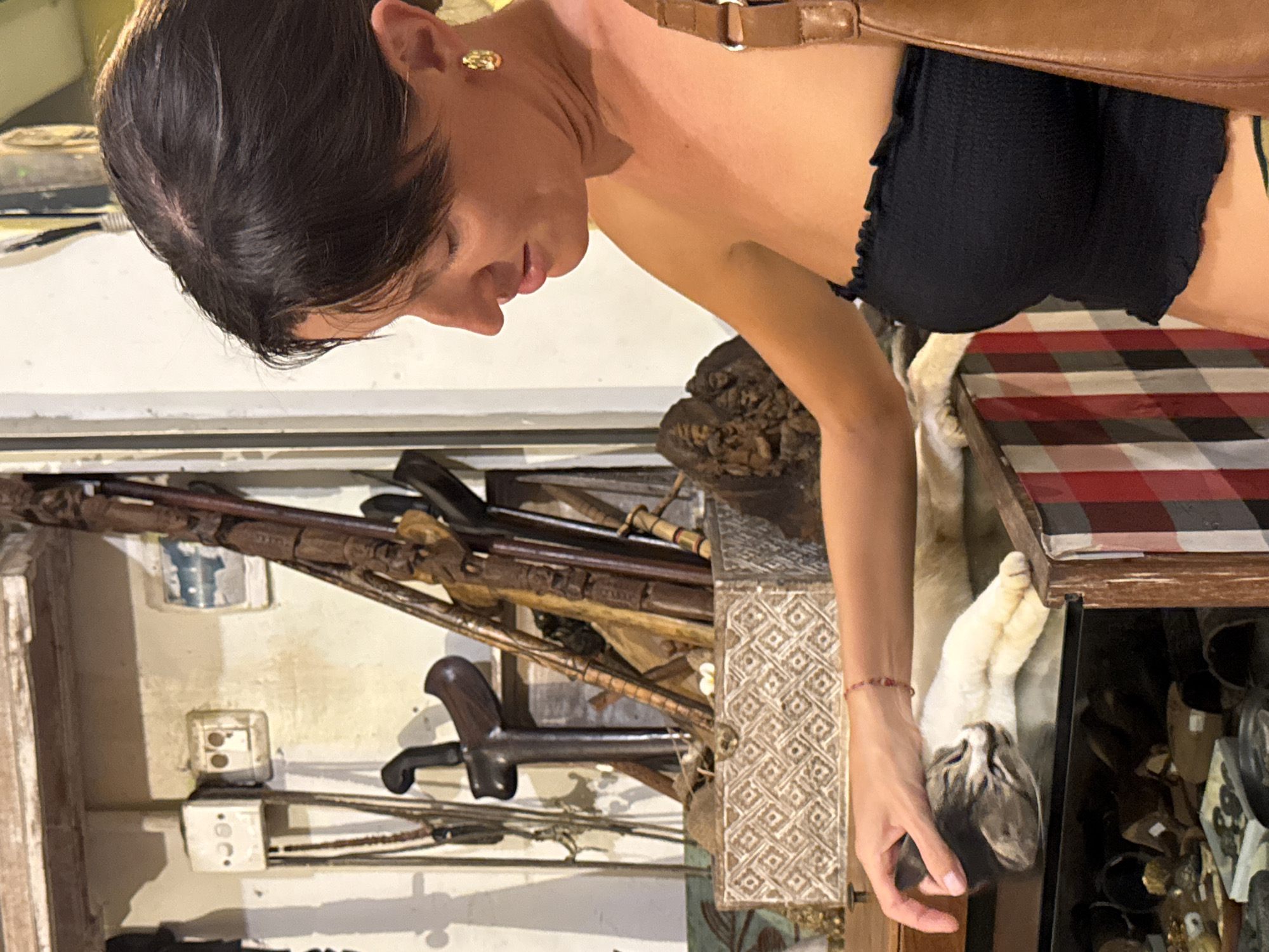 A woman wearing a black top and gold earrings is seen indoors, likely in a shop in Kuta Selatan, Indonesia. She is bending over and looking down, touching a display case. Within the display case are various objects, including wooden carvings, possibly weapons or tools, and electrical outlets. A white cat with grey markings is resting inside a drawer of the display case, which has a checkered pattern. Other items, possibly for sale, are visible in the shadowed areas of the case. The woman has a brown strap over her shoulder.