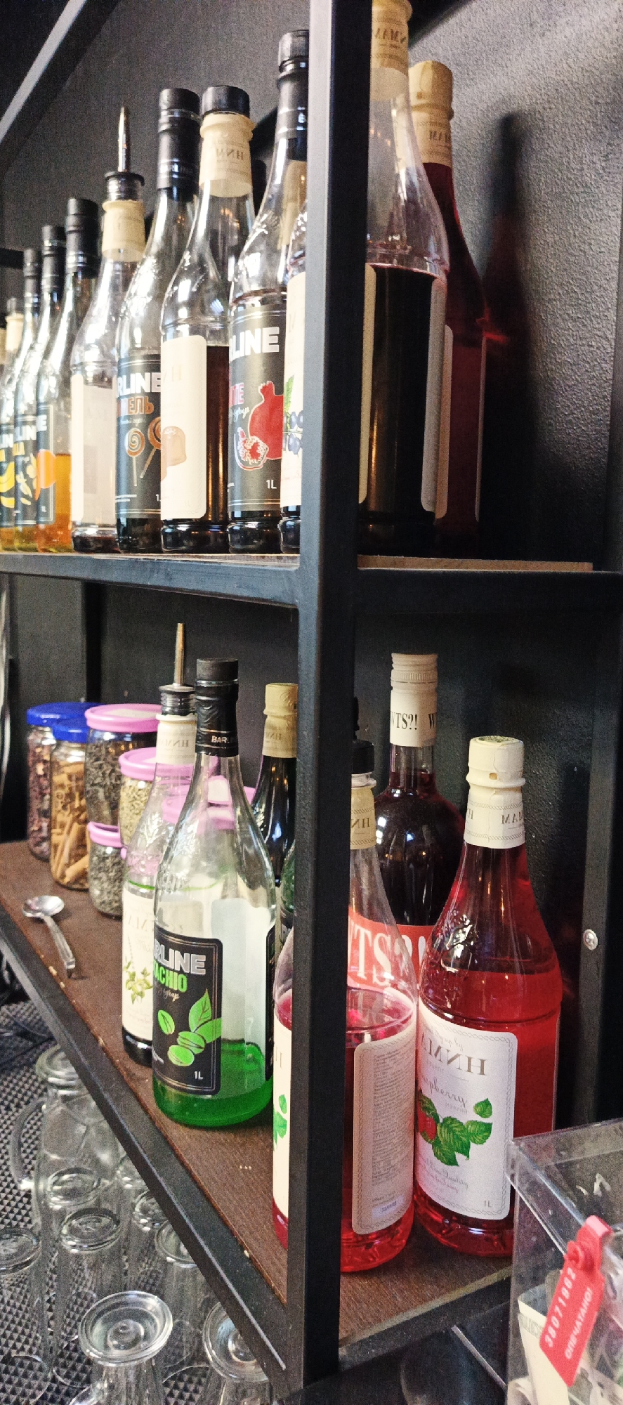 The image shows a shelf in what appears to be a bar or cafe. On the top shelf, there are several bottles of syrups or liqueurs, some with colorful labels. On the bottom shelf, there are jars containing dry goods, more bottles of syrups, and a clear glass pitcher with several glasses. The shelves are made of dark metal, and the background is a textured gray wall. The overall scene is well-lit and organized, suggesting a professional establishment. There are no people visible in the image. The presence of syrups and glassware indicates that this is a place where beverages are prepared and served. Based on the context of previous images, this scene is located in Votkinsk, Russia.