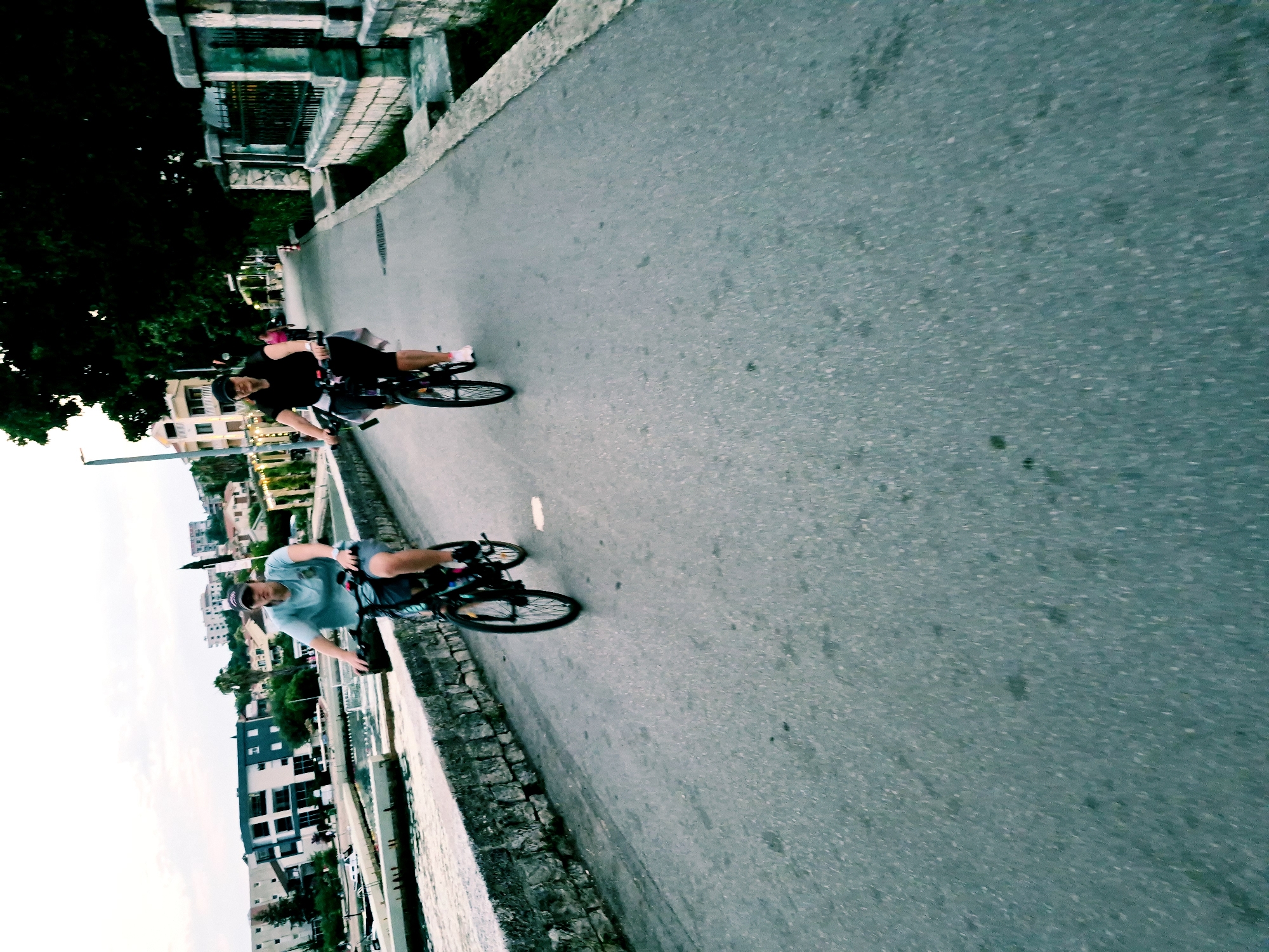 Two people are riding bicycles along a paved path beside a body of water, likely a bay or river, as suggested by the surrounding buildings and vegetation. The path is bordered by a stone wall on the water side and a concrete barrier on the land side, which is lined with trees and buildings. In the background, there are several multi-story buildings with tiled roofs, typical of Mediterranean architecture.
The activity is cycling, with both individuals appearing to be in motion. The person in the foreground, wearing a light blue t-shirt and shorts, is looking towards the camera and has a bicycle with black tires and a visible chain. The person in the background is wearing a black t-shirt and shorts and is also on a bicycle. The lighting suggests it is either early morning or late afternoon, with diffused light and a generally overcast or hazy sky. The overall mood seems relaxed and active, as they are enjoying a bike ride in an urban coastal environment. No visible text is present in the image. The crops suggest that the location is Đenovići, Montenegro.