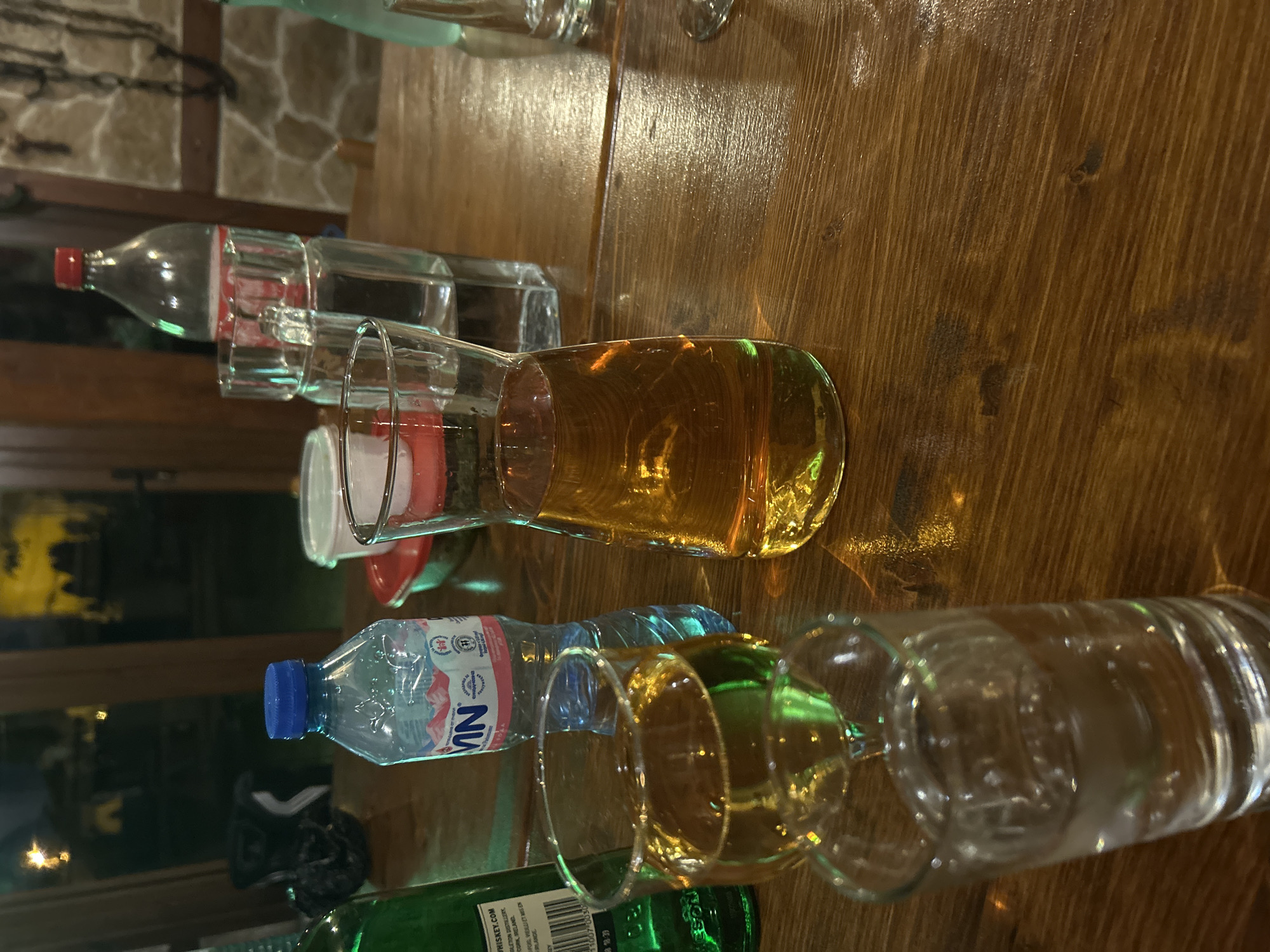 A close-up, slightly high-angle, medium shot shows a wooden table with several bottles and glasses on it. The bottles appear to be beverages, possibly water and alcohol. One bottle has a red cap, another a blue cap. There are also green bottles. The glasses contain different liquids, some clear and some amber in color.
The text "WHISKEY.COM" and "MN" are visible on the bottles. The "MN" bottle also features a logo that looks like a mountain range. A label indicates "Ireland" and "Mis En Rlande".
The setting appears to be indoors, possibly a restaurant or bar, with a stone wall visible in the background. The wood grain of the table is prominent. The location is provided as Mirovyane, Bulgaria.