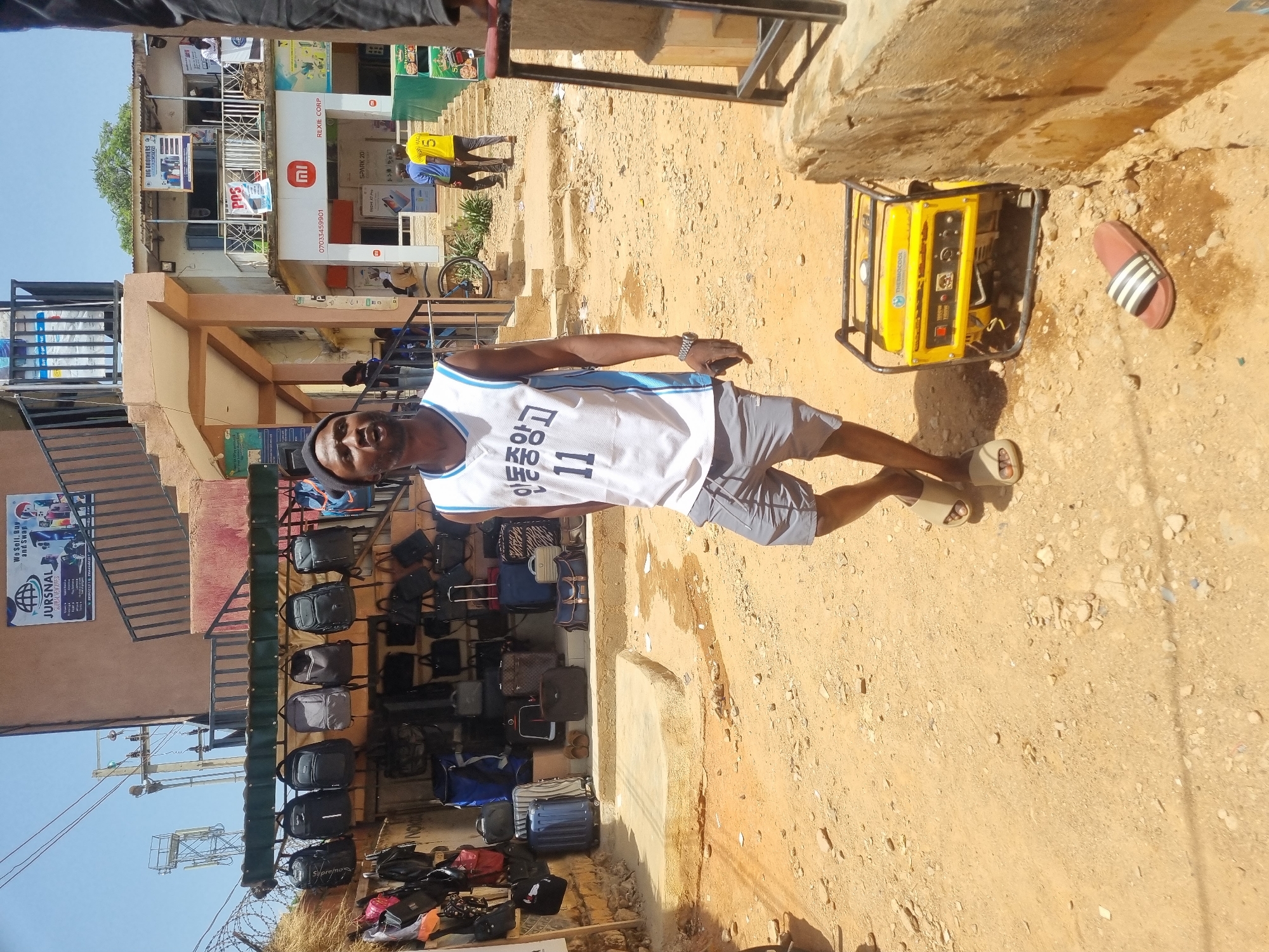 The image shows a man standing on a dirt road in Jos, Nigeria. He is wearing a white basketball jersey with the number 11 and Korean text on it, along with gray shorts and sandals. He has a watch on his left wrist.
To his right is a yellow Thermocool generator and a striped sandal. Behind him is a shop selling bags and luggage, displayed on a rack. Further in the background are buildings, including one with a Xiaomi advertisement and another with a sign for "BIG BROTHERS". There are also other people and a bicycle visible in the background.