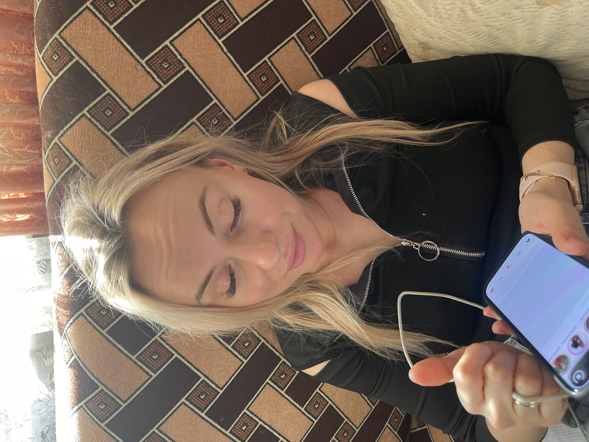 A woman with long blonde hair is reclining on a patterned brown and tan sofa. She is wearing a black top with a zipper. In her hands, she holds a smartphone and a white charging cable. The background appears to be an interior setting with patterned fabric. The city of Podolsk, Russia, cannot be determined from the given image.