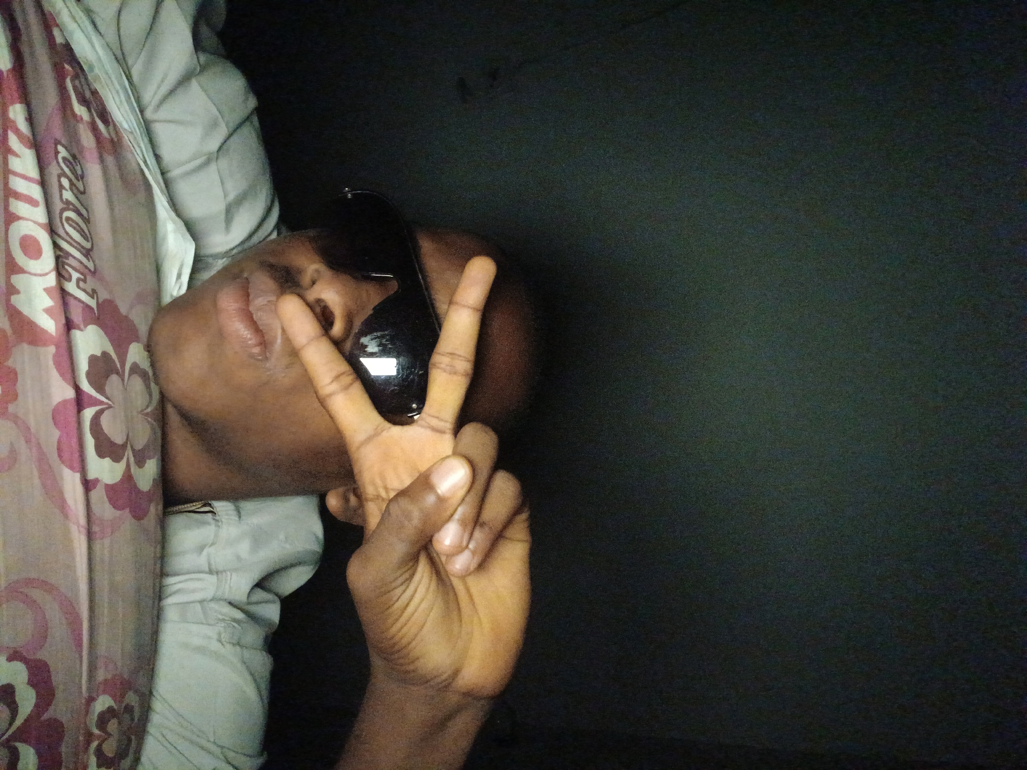 A medium shot captures a person wearing dark sunglasses and a light-colored collared shirt. The person is holding up two fingers in a peace sign near their face. They are leaning on what appears to be a bed or mattress with a floral pattern. The background is dark and nondescript. The lighting is dim, suggesting the photo was taken indoors at night or in a low-light environment. The image is grainy.