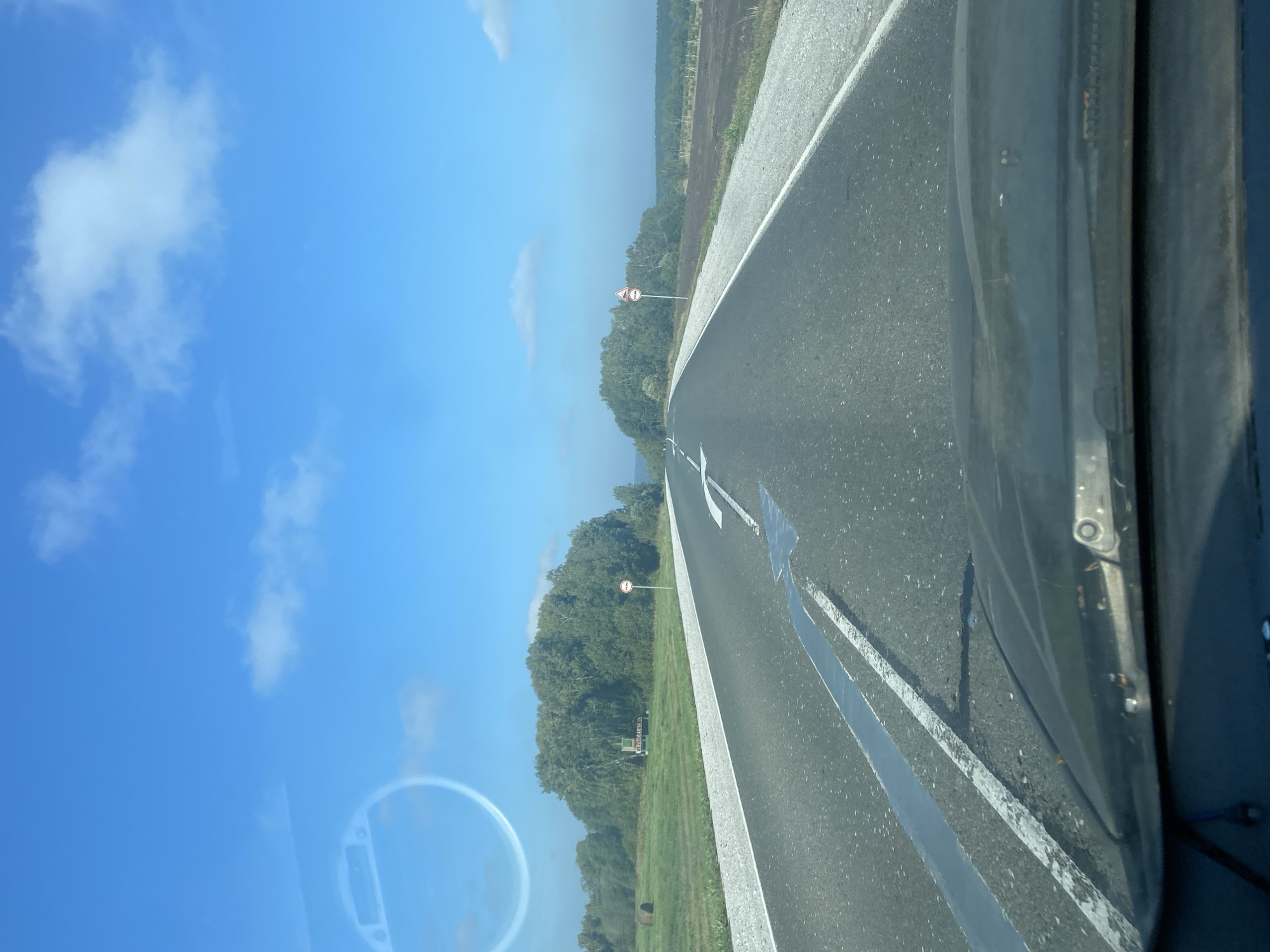 The image shows a first-person perspective from inside a vehicle traveling on a two-lane asphalt road under a bright blue sky with scattered white clouds. The road has white lane markings, with a large arrow indicating a turn or lane change ahead. On the left side of the road, there is a green field with a large bale of hay and a sign with text that appears to be in Russian. Further down the road on the left is a wooded area. On the right side of the road, the terrain appears to be a dark, cultivated field. There are two red and white circular traffic signs visible on poles along the roadside, one on each side. The dashboard of the vehicle is visible at the bottom of the image. The environment suggests a rural or semi-rural setting outside of the city of Podonino, Russia.