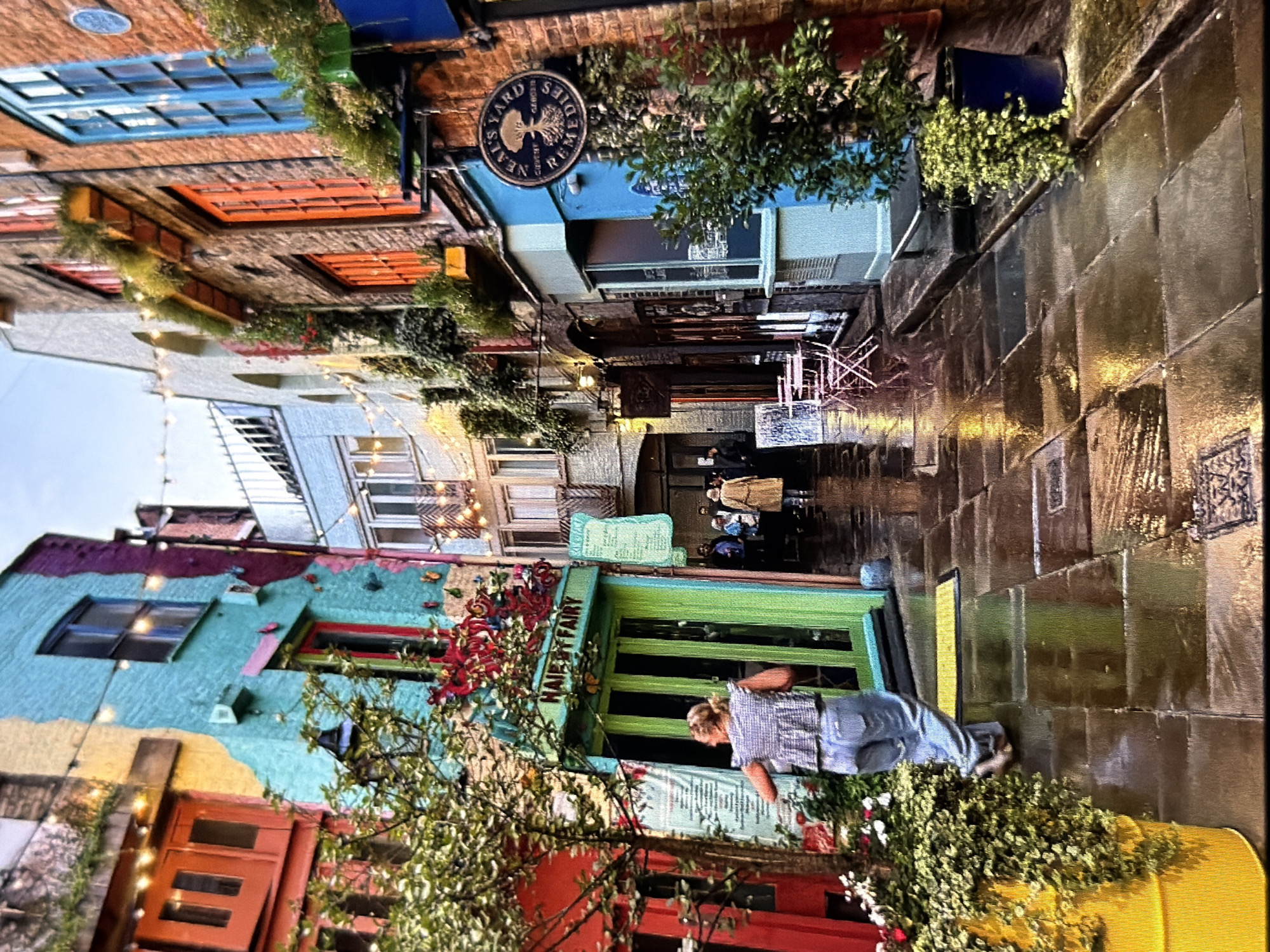 This image captures a narrow, wet cobblestone alleyway in Maidstone, United Kingdom, lined with multi-colored buildings under an overcast sky, suggesting late afternoon or early evening.
On the left side, a vibrant red building is partially obscured by a large yellow planter overflowing with green foliage and red flowers. Adjacent to it, a building features a green storefront with "HAIR & FAIRY" written above the door, which is adorned with red decorative elements. This building's upper facade is painted in sections of yellow, teal, and purple, with a dark window on the teal section. A single blonde woman, wearing a checkered top and light blue jeans, walks away from the viewer towards the green shop, her left arm slightly extended. A small potted tree stands to her left.
On the right side, brick buildings with contrasting window frames (some orange, some blue) line the alley. A prominent light blue storefront features a circular sign reading "NEAL'S YARD COVENT GARDEN REMEDIES" with a tree logo. A dark blue planter with green foliage stands below this sign. Further down the alley on the right, two pink metal chairs and a small table are visible outside a shop. String lights with warm bulbs are suspended across the alley at multiple points, illuminating the wet, reflective flagstone ground.
In the mid-ground distance, several indistinct figures are visible, including one person in a light-colored coat walking away from the viewer. The wet ground reflects the building colors and string lights, enhancing the illumination.