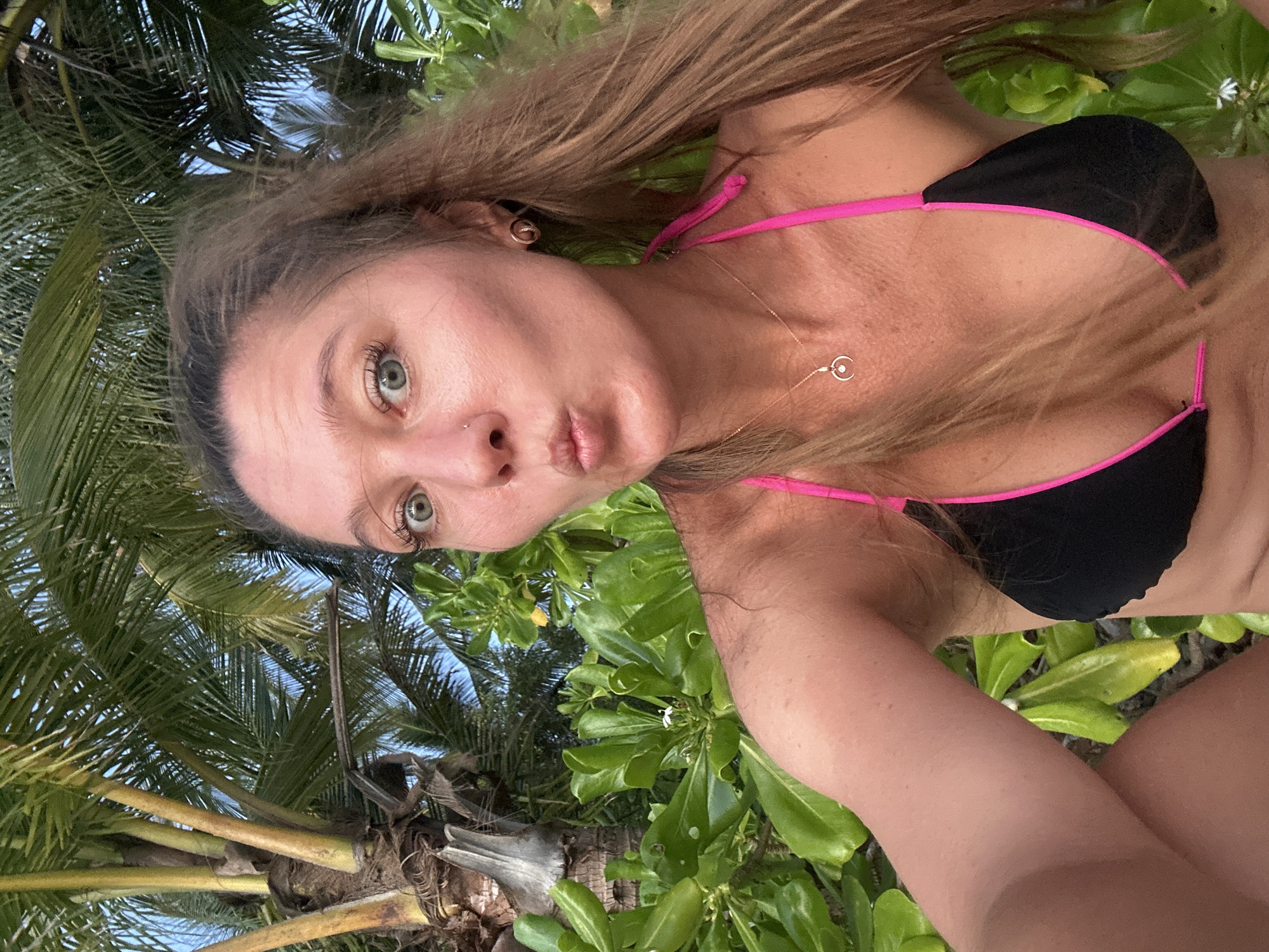 In Baan Taling Ngam, Thailand, a woman is captured in a selfie. She is wearing a black bikini with pink trim and a delicate necklace with a crescent moon pendant. Her hair is long and light brown, and she has green eyes. She is puckering her lips and looking at the camera. Behind her, lush green foliage and palm trees create a tropical backdrop, suggesting a beachside or garden setting. The lighting indicates it is either early morning or late afternoon.