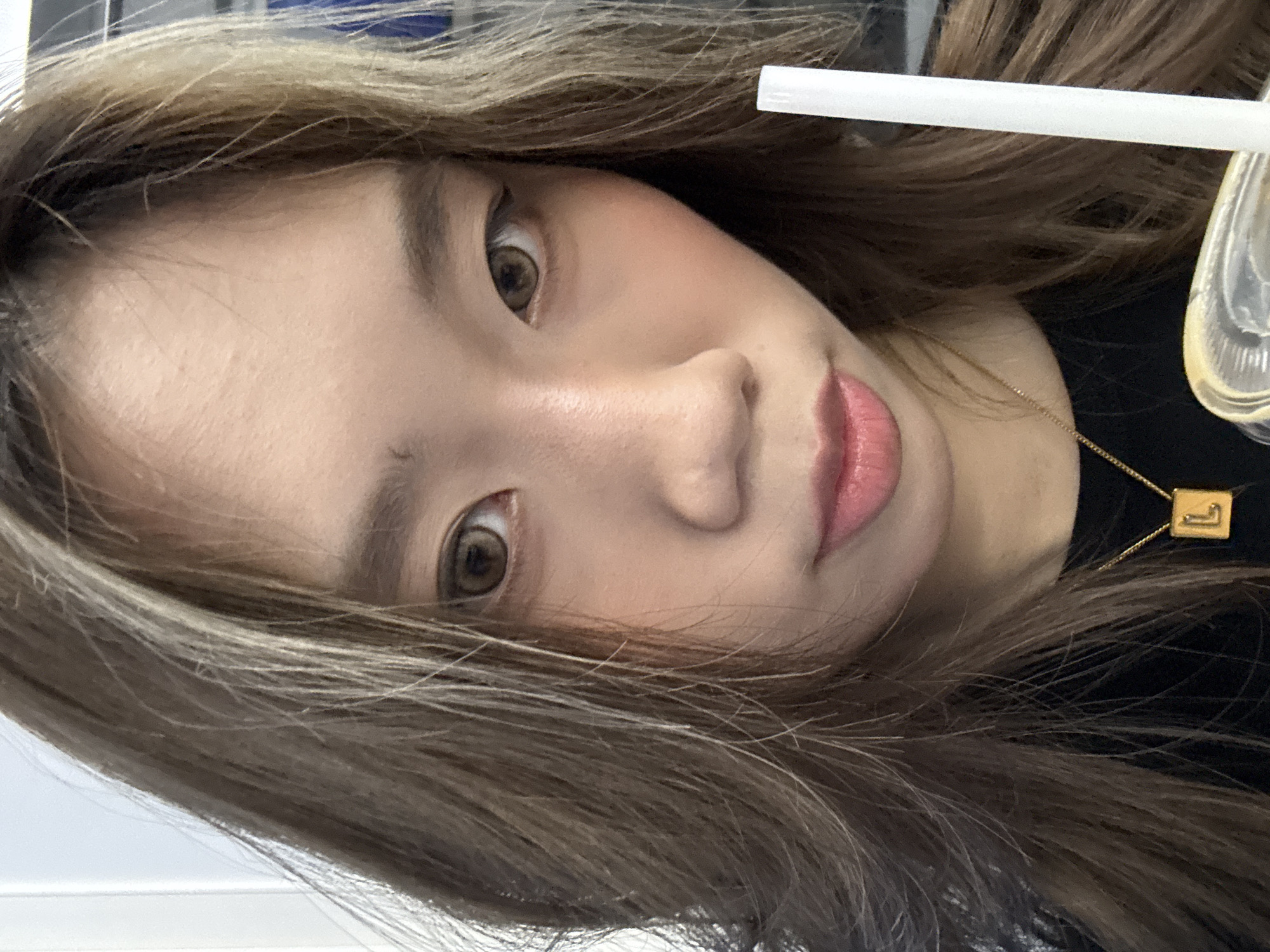 A close-up, tilted selfie of a young woman with light brown hair and eyes. She is wearing a black shirt and a gold necklace. A white straw is visible in the upper right corner. The location cannot be determined from the image.