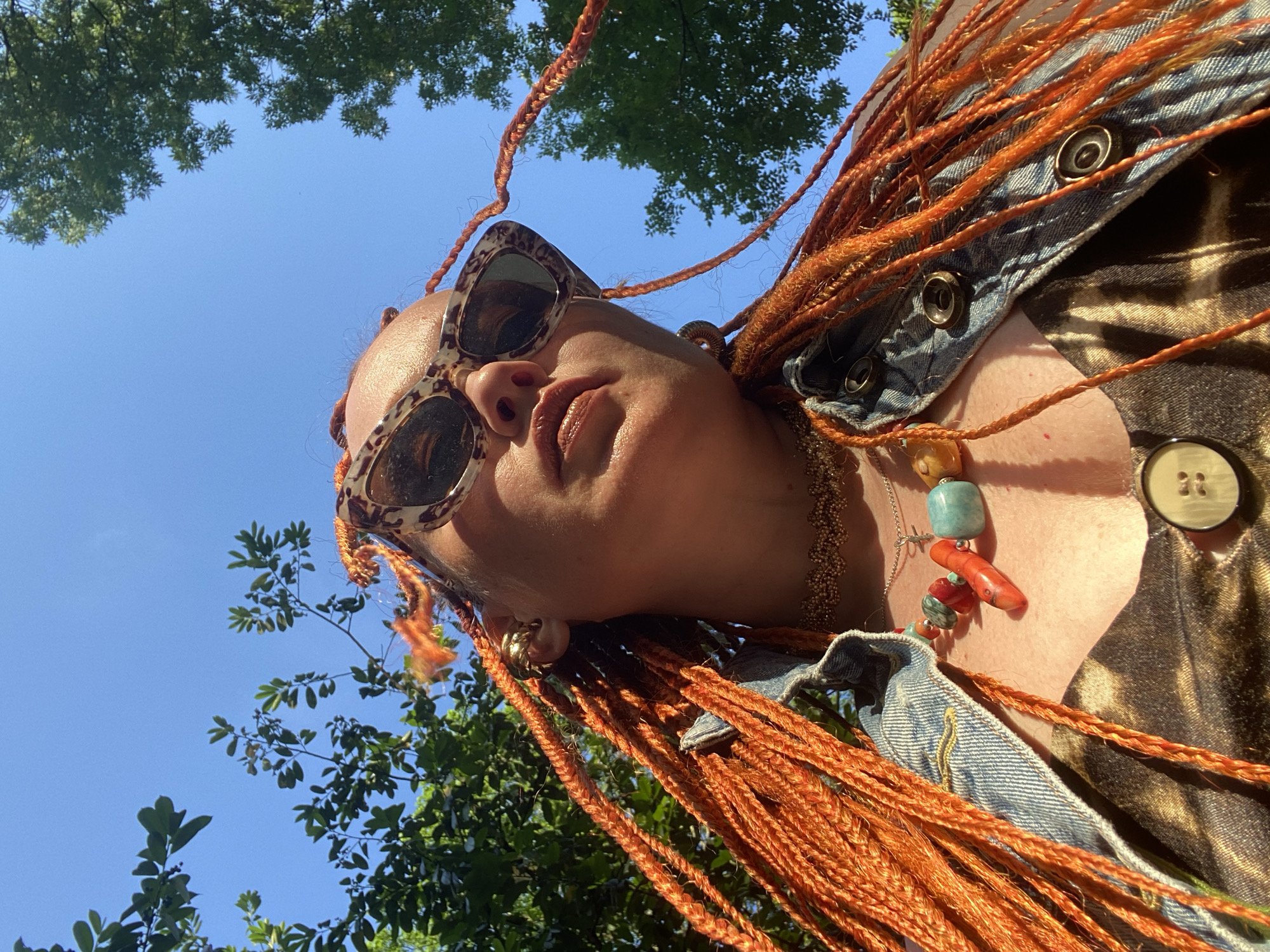 In an outdoor setting, likely a park or garden in Sochi, Russia, the image is a low-angle shot of a person wearing bright orange braided hair, sunglasses with a leopard print frame, and a denim jacket over a patterned top. The person is adorned with multiple necklaces, including one with large, colorful beads and another with a delicate chain. Sunlight illuminates the scene, casting shadows and highlighting the textures of the clothing and hair. The background consists of a clear blue sky and lush green foliage from trees. The overall impression is a candid, sun-drenched portrait.