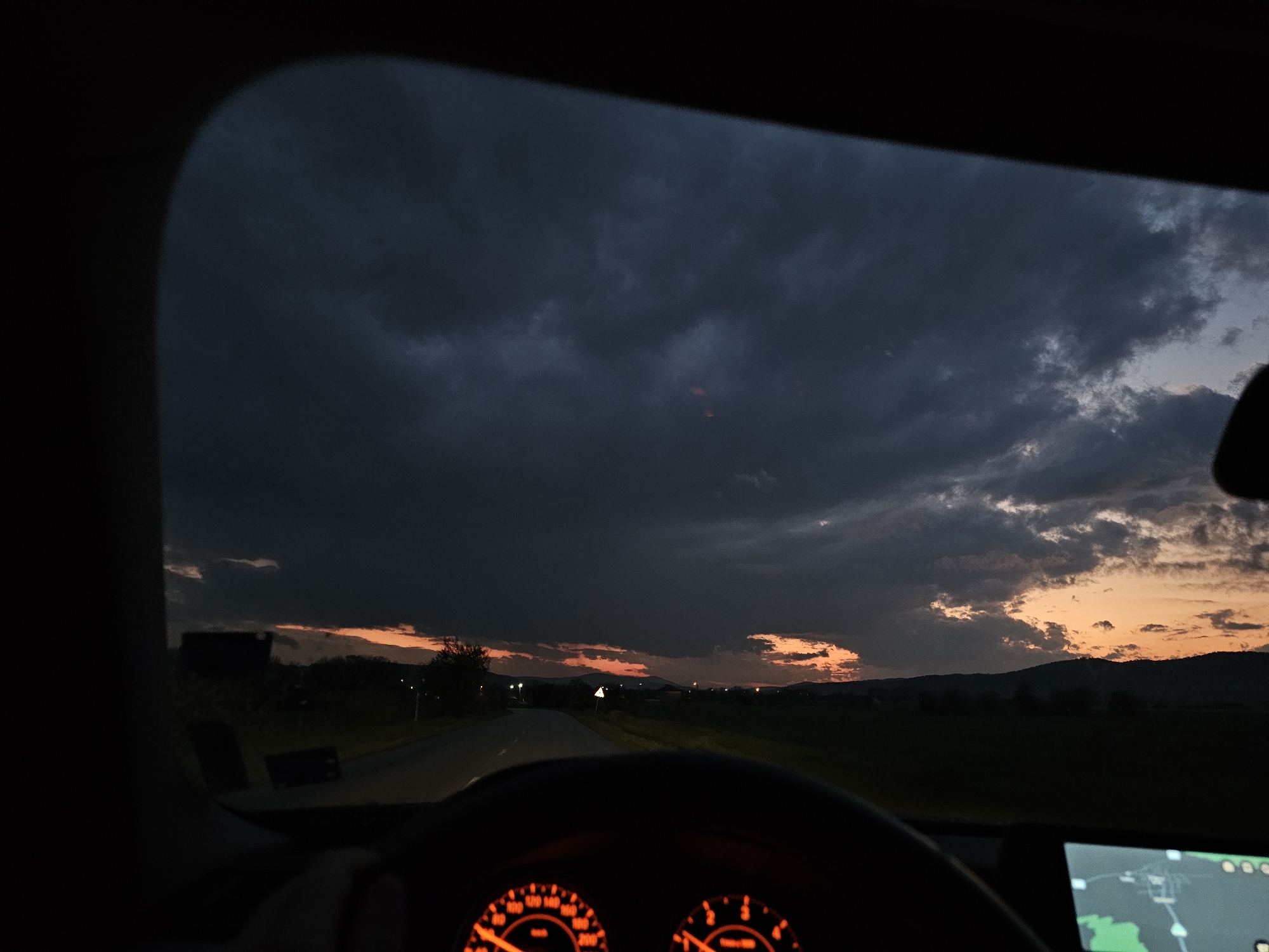 The image shows a view from inside a car, likely the driver's seat. The car is moving on a road outside the city of Krasnovo, Bulgaria, as indicated by the navigation system. The dashboard and steering wheel are visible, with illuminated gauges. Outside the windshield, a road stretches into the distance under a dramatic sky of dark, heavy clouds mixed with the orange glow of a sunset. The landscape on either side of the road consists of fields and distant hills or mountains. The scene appears to be at dusk or dawn, given the limited light. There are no people visible in the image.