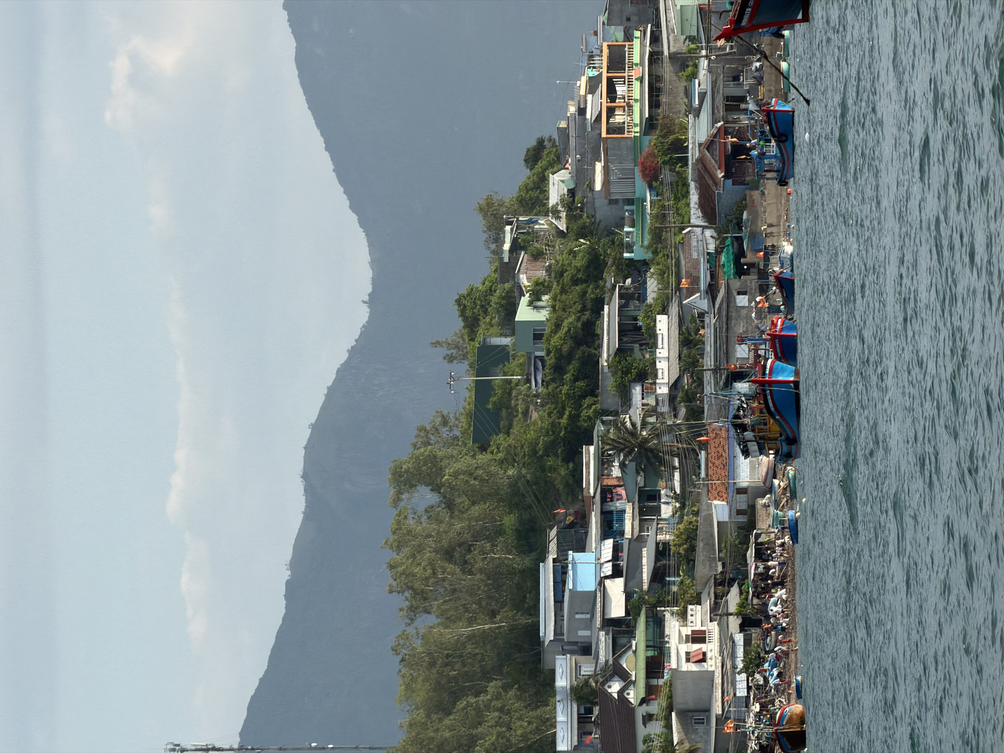 The image captures a coastal settlement in Nam Nha Trang Ward, Vietnam, viewed from a body of water. The foreground displays choppy blue-green water with numerous fishing vessels. Prominently, several large wooden fishing boats, predominantly painted in blue and red, are anchored or moving; one is distinctly labeled "AQUMAN." Smaller, light blue boats are scattered closer to the shoreline. On the sandy beach, a concentration of people is observed, engaged in various unspecified activities, likely related to the local fishing economy. A person is visible operating a long pole from a small boat near the shore.
The midground reveals a village densely packed onto a sloping hillside. The architecture comprises an array of multi-story houses, varying in color and style, often featuring balconies and corrugated roofs. Specific notable buildings include a bright blue house, an orange-framed structure, and a green-walled residence. Power lines and utility poles are visible throughout the residential area. Flags, possibly national flags, fly from several buildings. Lush green trees and vegetation are interspersed among the dwellings and cover the upper reaches of the hillside.
The background is dominated by a range of large, dark blue-grey mountains that rise steeply, forming a natural barrier behind the village. The sky above the mountains is a light, hazy blue, with several scattered white clouds. On the far left periphery, a tall, metallic communications tower ascends, partially silhouetted against the sky and mountain peaks. The scene is illuminated by clear daylight.