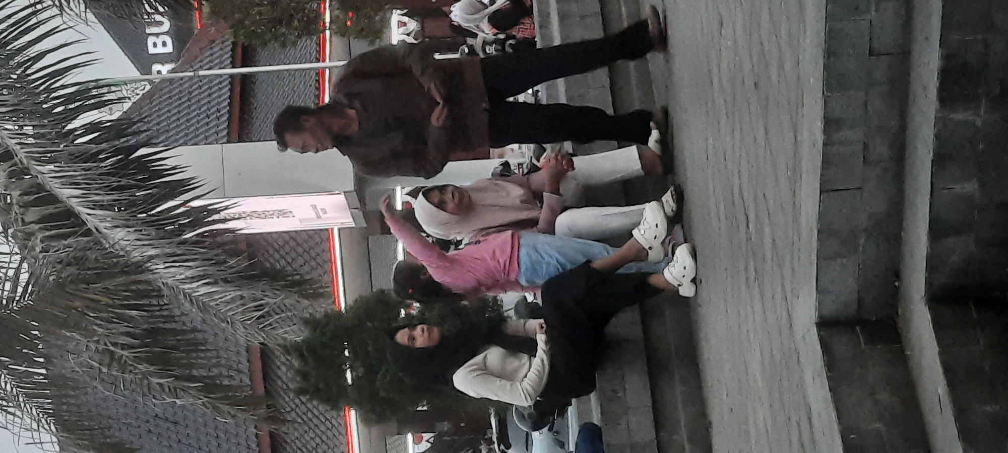 This image depicts a street scene in Kanigoro, Indonesia. In the foreground, four individuals are seated on a stone ledge, likely a public seating area. On the far left, a woman wearing a black hijab and a light-colored top is seated. Next to her, a young girl in a pink shirt and blue jeans is standing and reaching out her right arm. To her right, a woman in a light-colored hijab and pink top is seated, her hands clasped. Further to the right, a man in a brown jacket and dark pants stands, looking to his right. The background features a building with a dark, tiled roof and a sign that appears to say "R BUZ". Palm trees are visible overhead, suggesting a tropical or subtropical environment. The overall lighting suggests it may be dusk or early morning.
