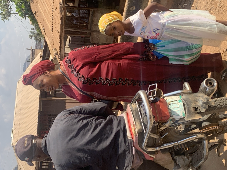 This image depicts an outdoor scene in Egbedi, Nigeria, under bright daylight. Three individuals are present on an unpaved, dirt ground.
In the foreground, a man is seated on a motorcycle, viewed from his back. He wears a dark, possibly black, long-sleeved shirt and a dark baseball cap with "TEAM JB" inscribed in white lettering on the back. The motorcycle has a red and white seat, a chrome rear rack, and a visible rear tire with a chain and an exhaust pipe. A partially obscured, light blue/green license plate is affixed to the rear fender, which appears worn.
To the right of the motorcycle and the man, a woman stands, facing slightly towards the left of the frame. She wears a long, deep red dress adorned with black, leaf-like embroidery down the front and along the sleeves. Her head is covered with a matching red headwrap. She carries a dark shoulder bag. Her mouth is slightly open, suggesting she is speaking or in mid-sentence.
Standing to the woman's right is a young girl. She wears a white short-sleeved t-shirt featuring a colorful graphic, likely of animated ponies. Her lower half is covered by a light-colored, possibly multi-colored, ruffled skirt. A yellow headwrap covers her hair. She holds two striped plastic shopping bags in her left hand and looks directly towards the viewer.
The background features several structures with corrugated metal roofs and simple wall construction, characteristic of local architecture. Distant hills or mountains are visible under a clear, bright sky.
