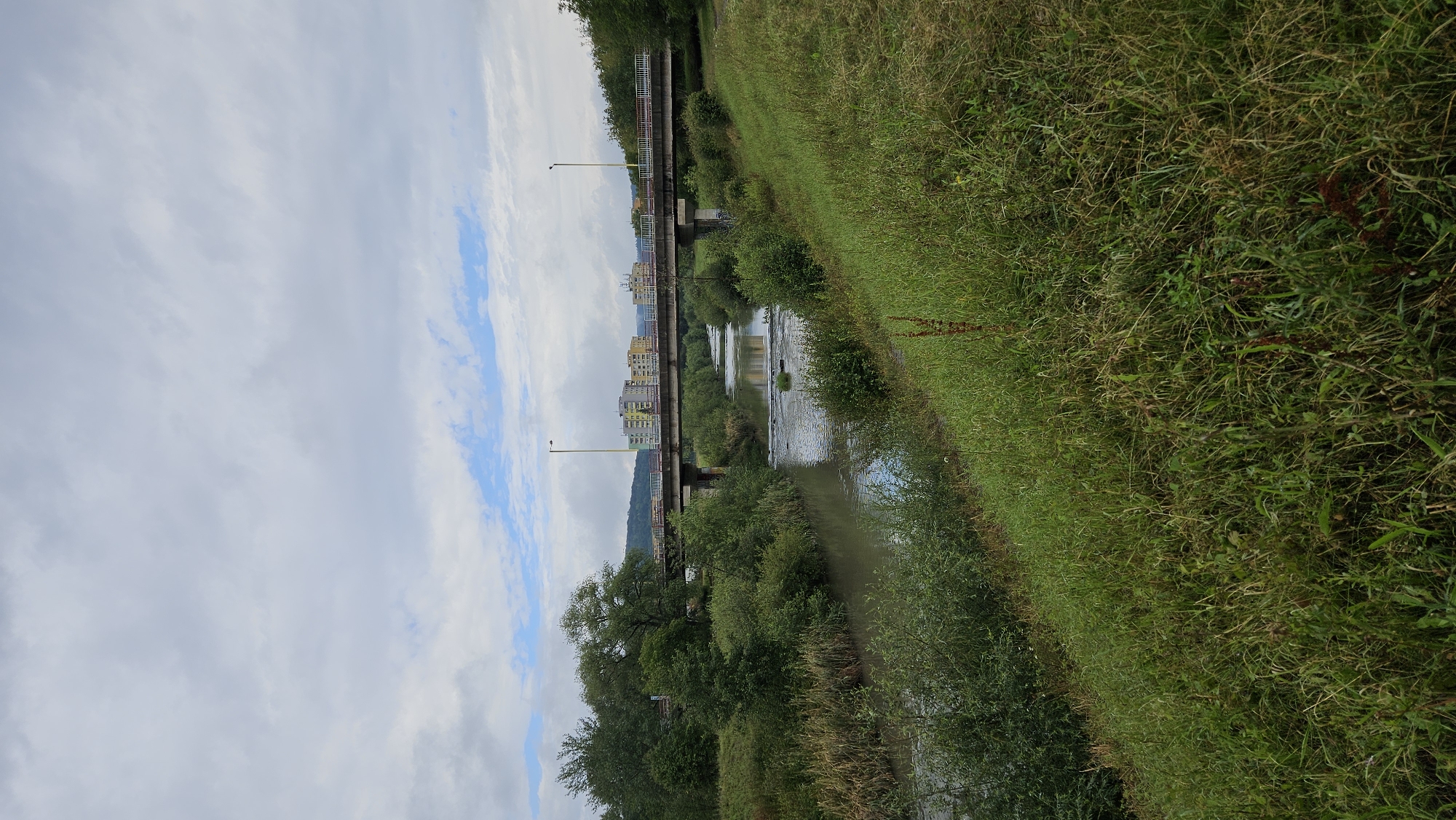 The image depicts a wide shot of a river flowing through a somewhat urbanized area, likely in Prešov, Slovakia. A concrete bridge with railings spans across the river in the mid-ground. On the far bank, several multi-story apartment buildings are visible, with some trees and vegetation interspersed. The immediate foreground and riverbanks are covered in lush green grass and dense bushes, suggesting a park or natural area bordering the river. The sky is overcast with patches of blue sky peeking through. The overall scene is calm and appears to be a day with moderate weather.