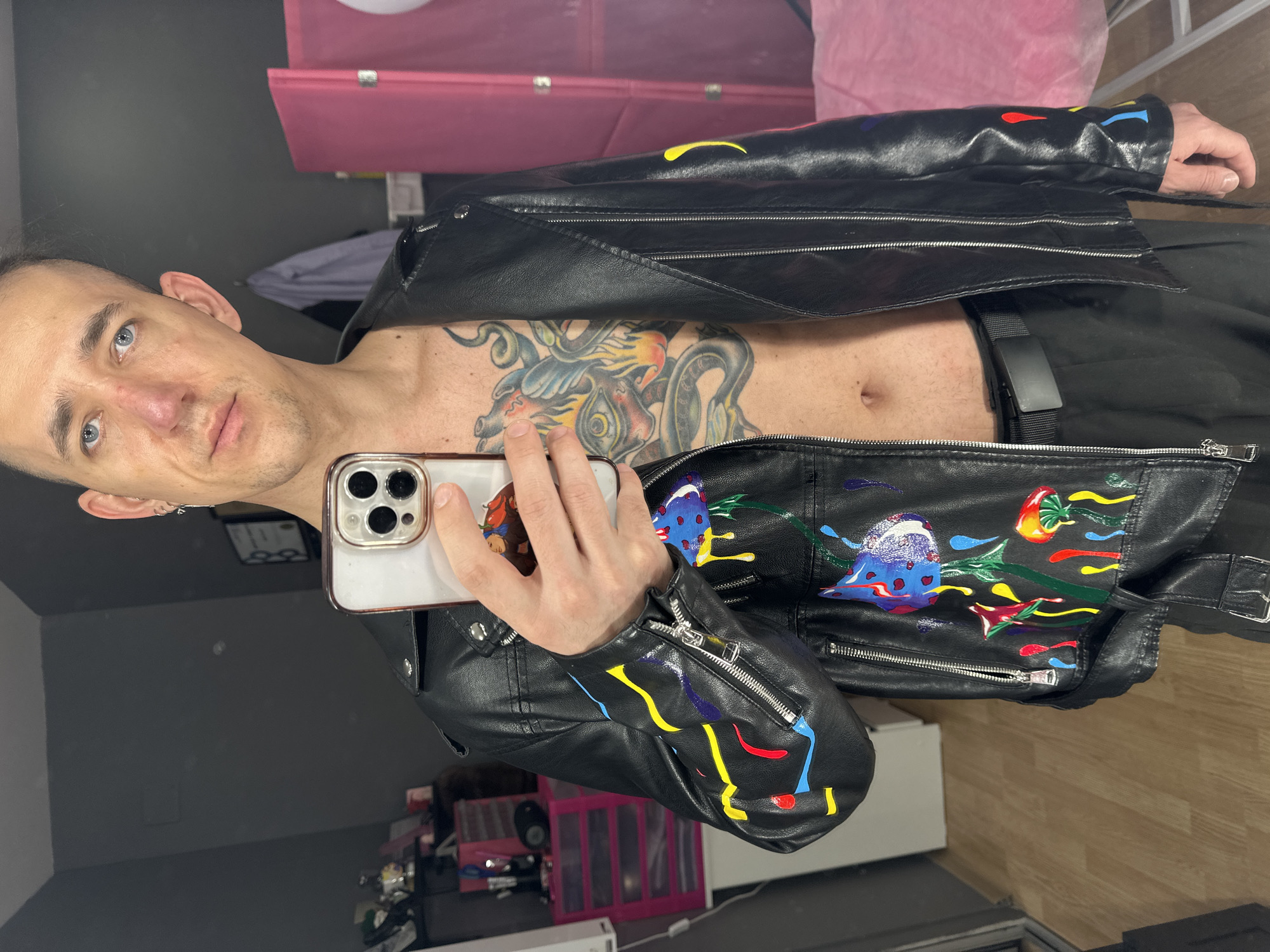 A fair-skinned person with short hair and light eyes is holding an iPhone and taking a selfie in what appears to be a room or dressing area with a grey wall and a wooden floor. The person is wearing a black leather jacket with colorful paint splatters, and a belt. A large tattoo is visible on the person's chest and stomach. Behind the person, there is a partially visible pink screen and some other items, including what looks like toys. There's no information to conclude this is in the city of Saratov, Russia.