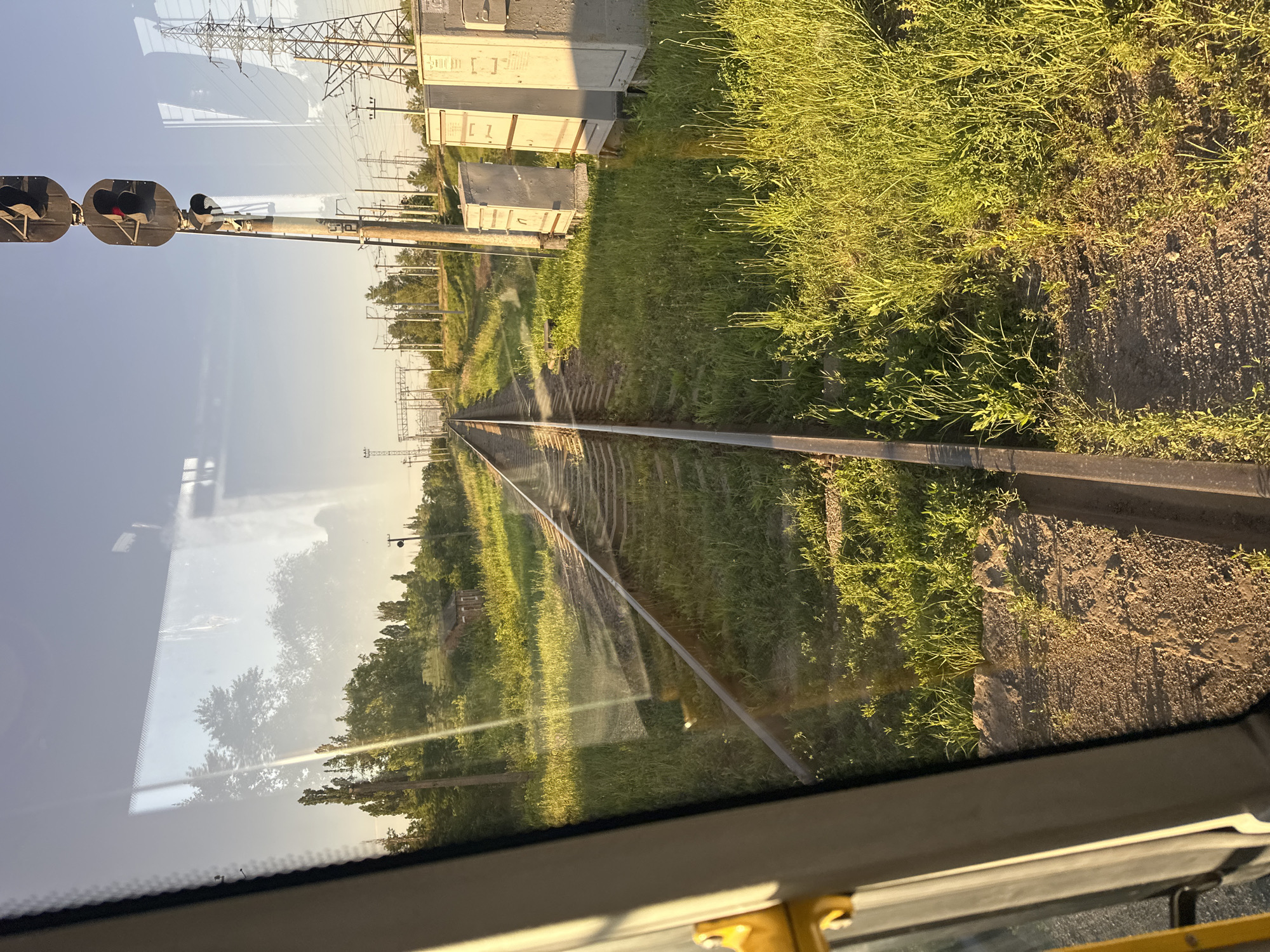 A view from inside a vehicle, likely a train, shows railroad tracks stretching into the distance under a clear sky. The sun casts long shadows, indicating it is either early morning or late afternoon. Green grass and trees line the tracks, with a small brick building visible among the trees on the left side. A railway signal with a red light is prominently displayed on the right, suggesting the train may be stopped or preparing to stop. Electrical transmission towers and utility boxes are also present along the tracks. The reflection on the vehicle's window shows trees and the sky. The scene is consistent with a rural or semi-rural area, possibly on the outskirts of a town like Dmytrivka, Ukraine.