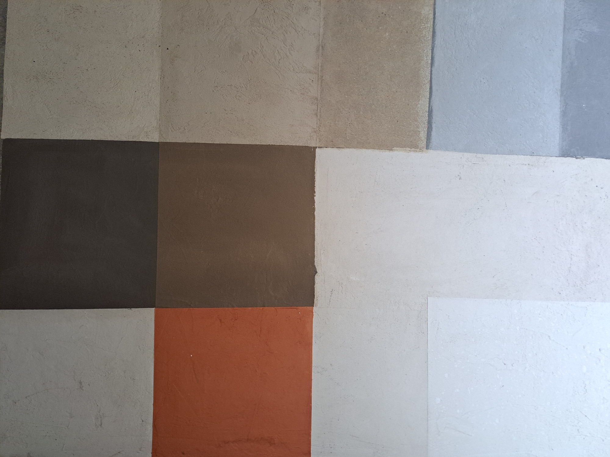The image shows a sample board with various paint colors and textures, likely intended for interior or exterior design purposes. There are nine distinct squares, each displaying a different shade and finish. Colors range from neutral tones (beige, gray, white) to bolder hues (brown, orange). The texture varies, with some squares appearing smooth and others showing a rougher, more stucco-like finish. The setting is not identifiable as being located in Yerevan, Armenia, based on the visual information provided. There are no people or actions depicted in the image.