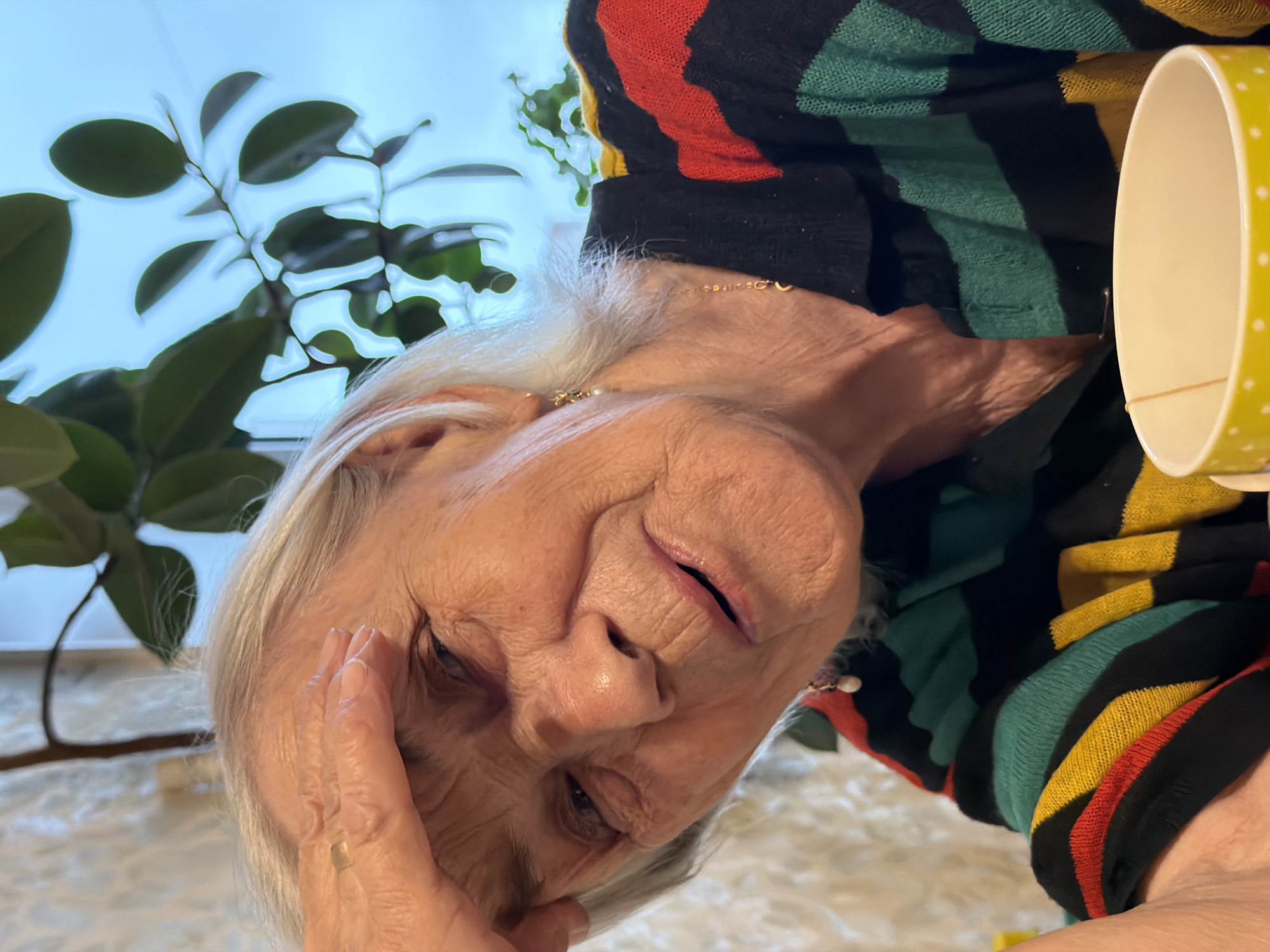 An older woman with gray hair and wrinkles, wearing a colorful striped sweater, is seen holding her head with her hand. She appears to be looking down. A yellow cup with white polka dots is visible near her shoulder. In the background, a plant with green leaves is present. There is no visible information to determine if the location is in Izhevsk, Russia.