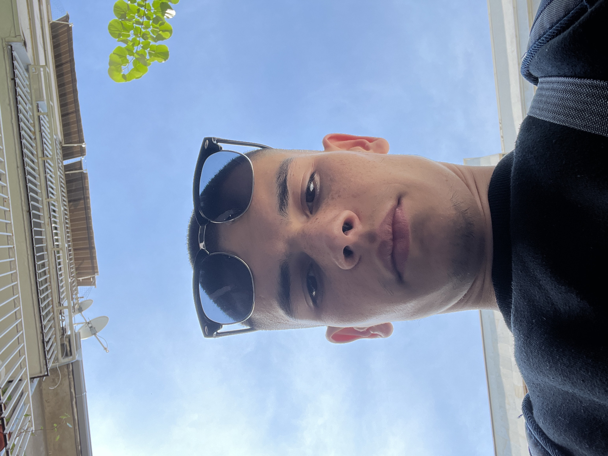 A low-angle, close-up shot shows a young man with short dark hair and sunglasses, looking down at the camera. The background reveals a light-colored building with balconies and a satellite dish, partially obscured by tree leaves, against a blue sky. The person is wearing a dark-colored garment. Based on architectural cues, the location could be Plovdiv, Bulgaria.
