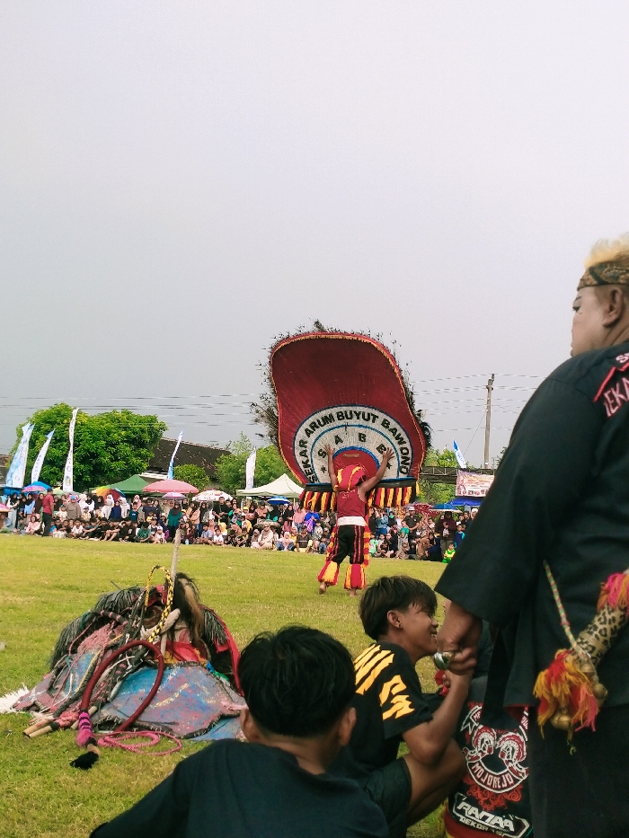 The image was taken in Karangmalang, Indonesia, at the coordinates -7.47 latitude and 111.064 longitude. The scene shows a performance of traditional Indonesian dance. A man in a red and yellow costume is performing a dance with a large, red, feathered headdress. The headdress is decorated with the words "Sekar Arum Buyut Bawono" and features feathers that extend out from the sides of the head. There are people in the background, watching the performance. The performer is holding a staff or cane in his right hand, and he appears to be leaping or spinning. The dancer is surrounded by a crowd of people sitting in a grassy area, watching the performance. Some of the audience members are wearing hats, scarves, and other accessories. The scene appears to be a celebration or festival. The day is partly cloudy, and the ground is covered in a thin layer of grass. To the right of the frame, an older man is wearing a black shirt with a large eagle design.