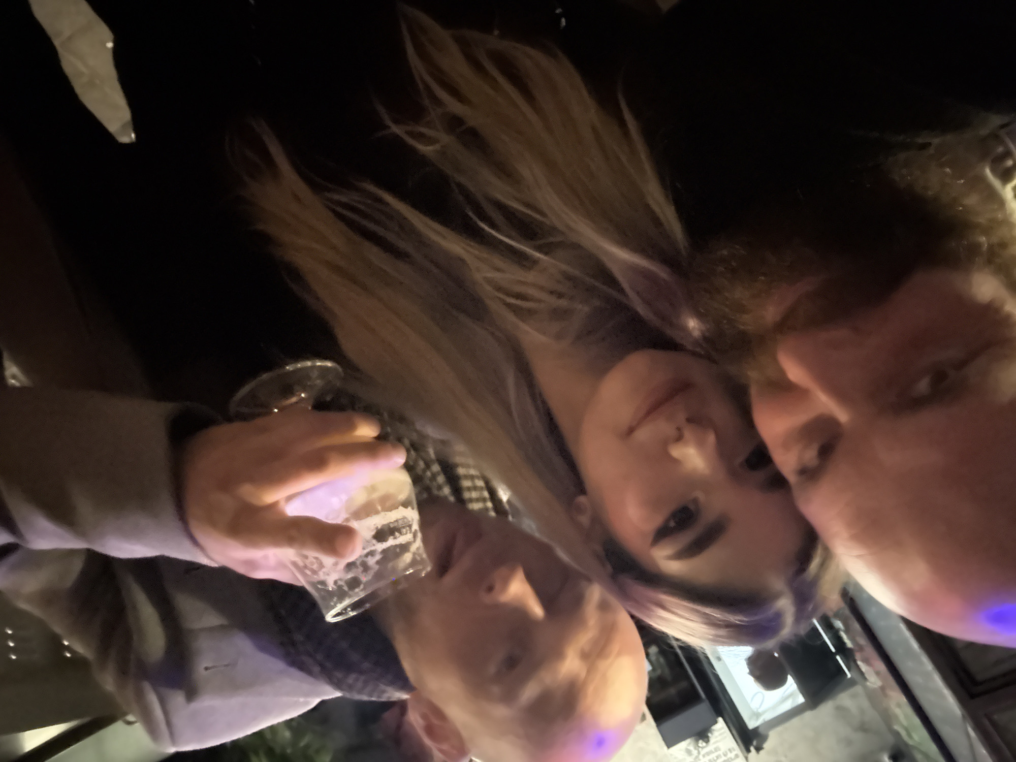The image depicts three individuals in a dimly lit indoor establishment, consistent with a bar or pub, located in Tel-Aviv, Israel.
In the left foreground, a bald adult male with a full brown beard is partially visible, facing towards the camera. In the center, a young adult female with long, light-colored hair, featuring subtle purple highlights, looks directly at the camera. She has distinct eyeliner and a small piercing on her right nostril. To the right, another bald adult male is seen holding a stemmed glass filled with a light, frothy beverage in his right hand. He wears a light-colored overcoat or jacket over a dark, possibly patterned, scarf and is looking slightly downwards. A faint, blurry reflection of a fourth bald individual is discernible in the deep background. The ambient lighting includes a noticeable purple hue illuminating the heads of the two foreground males. The background consists of dark, indistinct surfaces with some reflective elements, suggesting an enclosed commercial space.