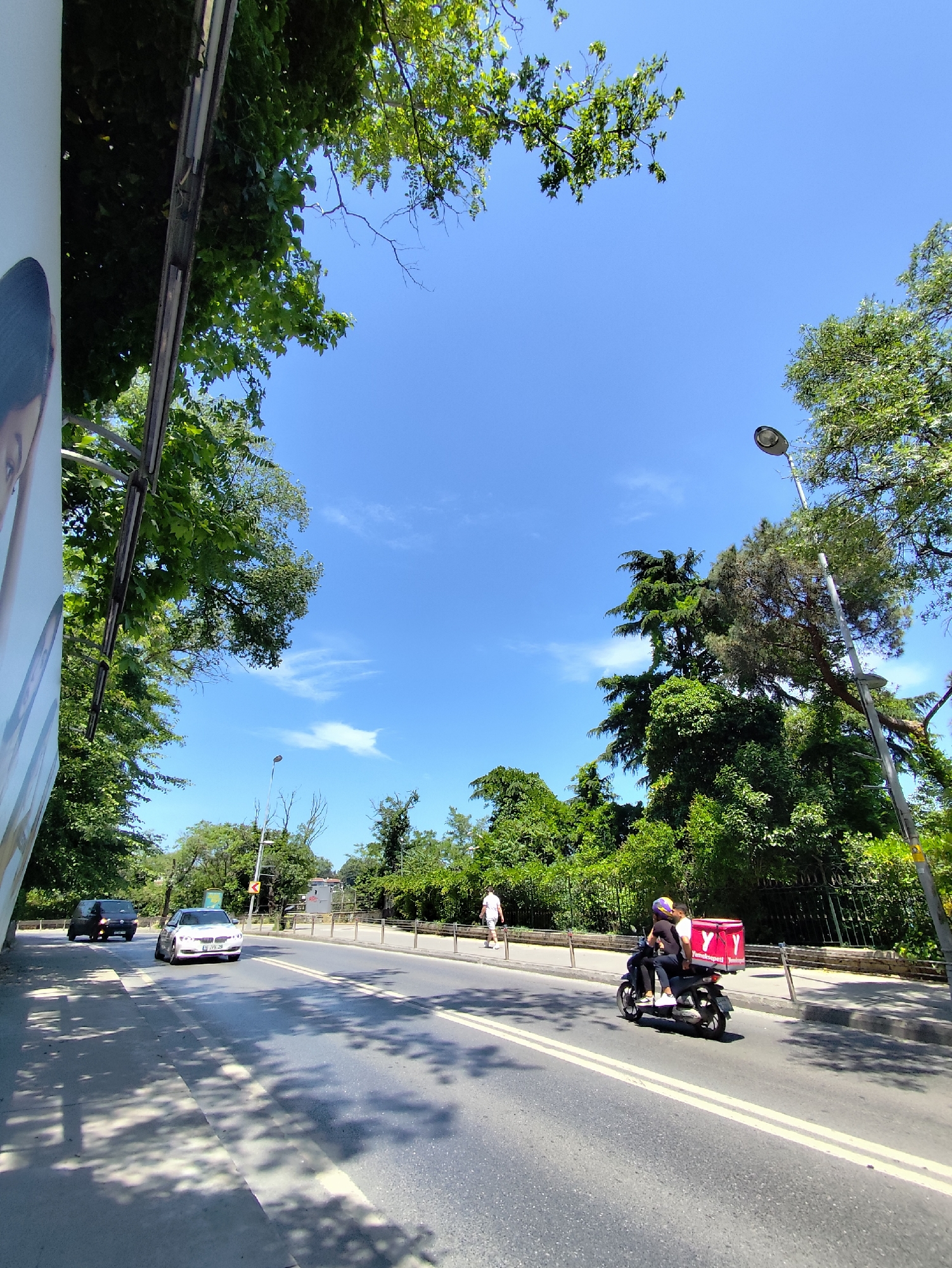 The image depicts a bright, sunny daytime scene on a road lined with lush green trees and foliage in Sarıyer, Turkey. The sky is a clear, vibrant blue with a few scattered, wispy white clouds. The perspective is slightly upward, showcasing the expanse of the sky and the dense canopy of trees framing the view.
In the foreground and midground, an asphalt road with double yellow lines is visible, marked by distinct shadows of trees. Several vehicles are present on the road. Two cars, a dark van and a white sedan, are seen further down the road, driving away from the viewer. Closer to the foreground, a black scooter with two riders is prominent. The driver wears a helmet, and a passenger is seated behind, holding onto the driver. Attached to the back of the scooter is a large, rectangular red delivery box featuring a white "Y" logo and the brand name "Yemeksepeti," indicating a food delivery service.
To the right of the road, a sidewalk is bordered by a metal barrier, separating it from the traffic. Beyond the barrier, a dense line of green trees and bushes extends, and a street lamp post is also visible. Further along the sidewalk, a pedestrian can be seen walking away from the viewer. On the far left, a portion of a large white billboard or banner structure is visible, showing part of a human face, suggesting an advertisement.
The scene captures a typical sunny day with ongoing traffic and daily activities, highlighted by the delivery scooter.