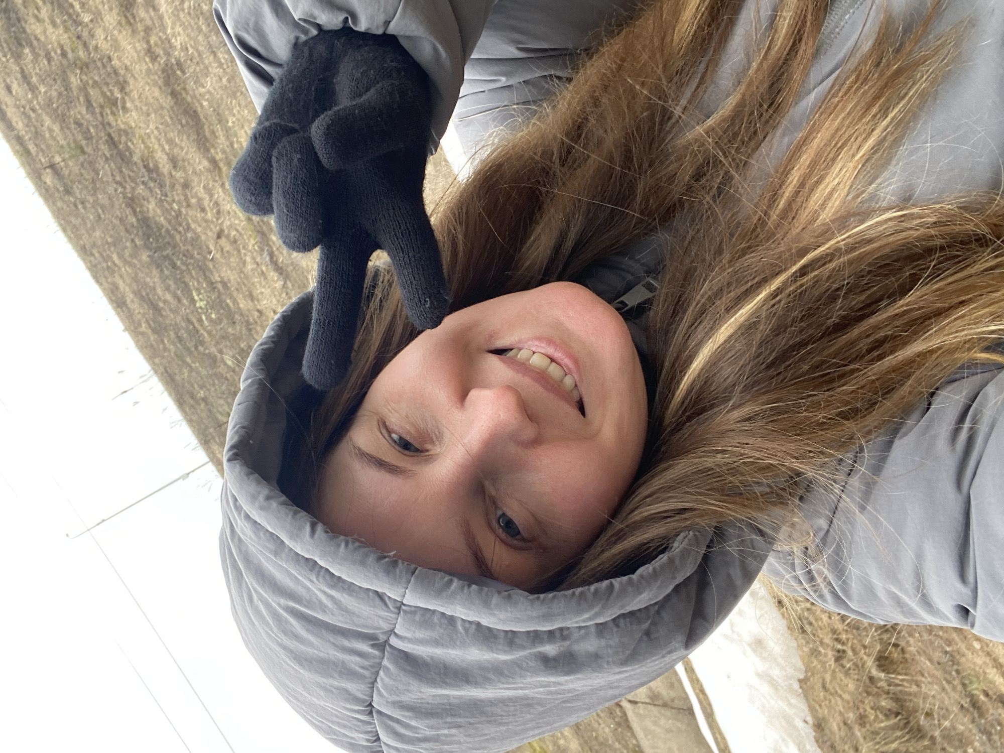 A woman with long, light brown hair and blue eyes is wearing a gray hooded jacket and black gloves. She is smiling and making a peace sign with her gloved hand. The background appears to be an outdoor scene with dry grass and a hint of white, possibly snow or a light-colored surface. The picture is rotated. The location is Noviy, Russia.