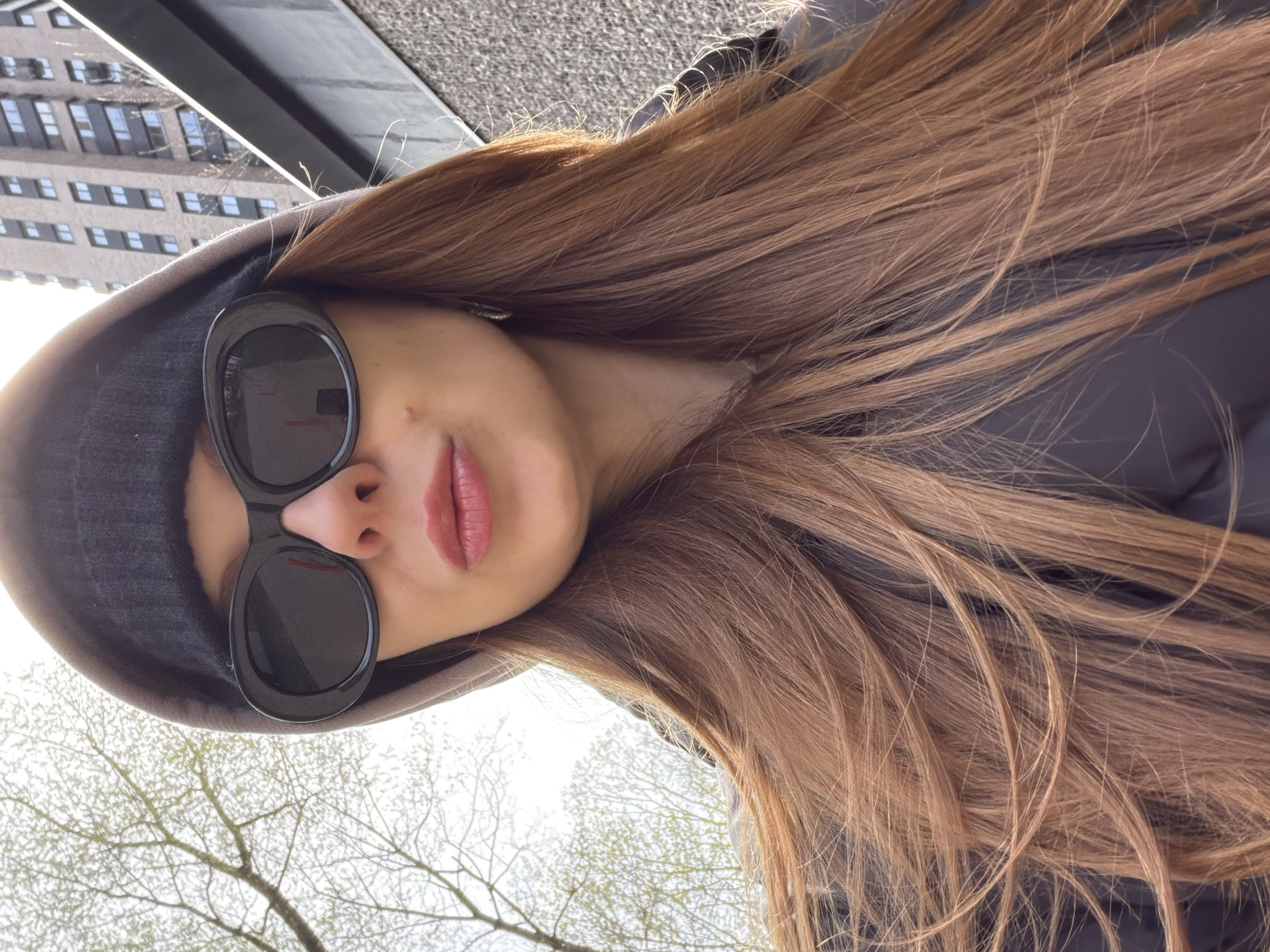 The image features a close-up, upward-angled portrait of a person, likely female, from the chest up, with her head tilted. She has long, straight to wavy light brown hair that cascades over her right shoulder and back. She is wearing a dark grey or black hooded top, with the hood pulled up around her head. Large, dark, oval-shaped sunglasses obscure her eyes, but her lower face is visible, showing a slightly parted mouth with pink or peachy lipstick and a small mole on the upper lip/philtrum area.
The background is divided. To the upper left, a modern building with numerous rectangular windows is partially visible under a bright sky, indicating an urban or suburban setting. To the lower left, bare tree branches with nascent buds or small leaves are silhouetted against a bright, possibly overcast, sky, suggesting late winter or early spring. The lighting is bright and natural, indicating daytime. The person appears to be posing for the camera. There is no visible text in the image. The location context for this scene is Khimki, Russia.