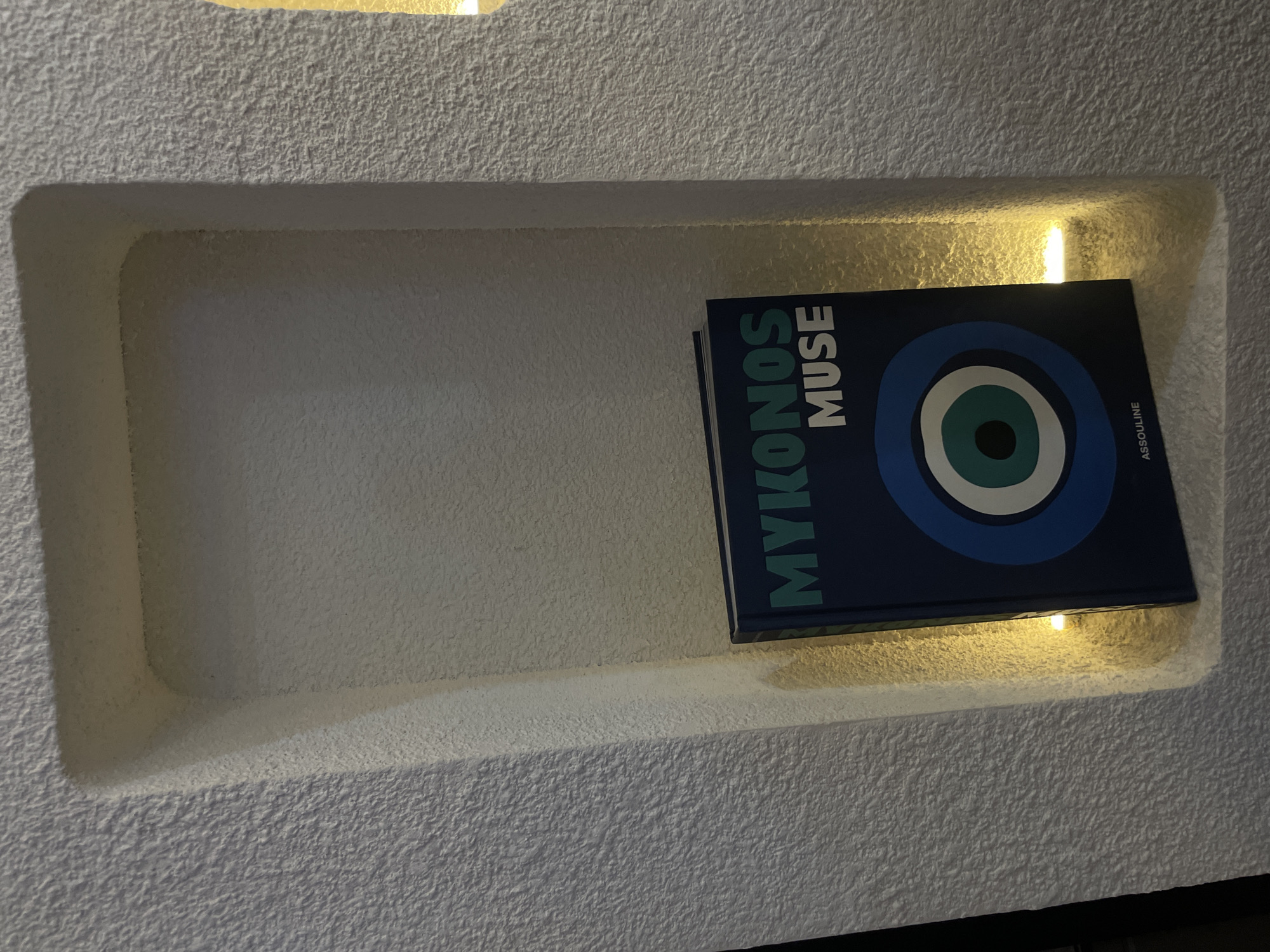 A blue book titled "Mykonos Muse" with a large blue and white circle design is placed in a recessed shelf in a white textured wall. The shelf is illuminated by a warm light source. The location is unknown and there is no information about Puebla, Mexico. There are no people or actions visible in the image.