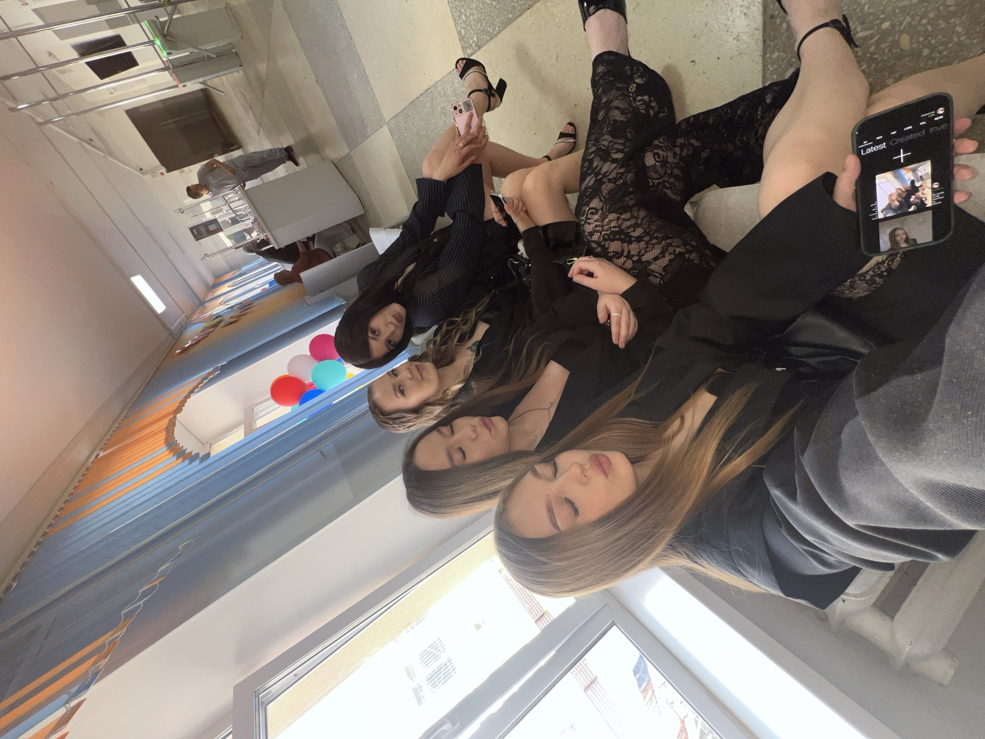 In Chaikovsky, Russia, four young women are seated on a bench in what appears to be a public building, possibly a school hallway. Two of the women in the foreground are leaning forward, with one holding a smartphone displaying photos. The other two women are seated behind them, with one looking towards the camera. The hallway is decorated with balloons and features a large window and tiled flooring. In the background, other individuals are visible, along with some office-like furniture.