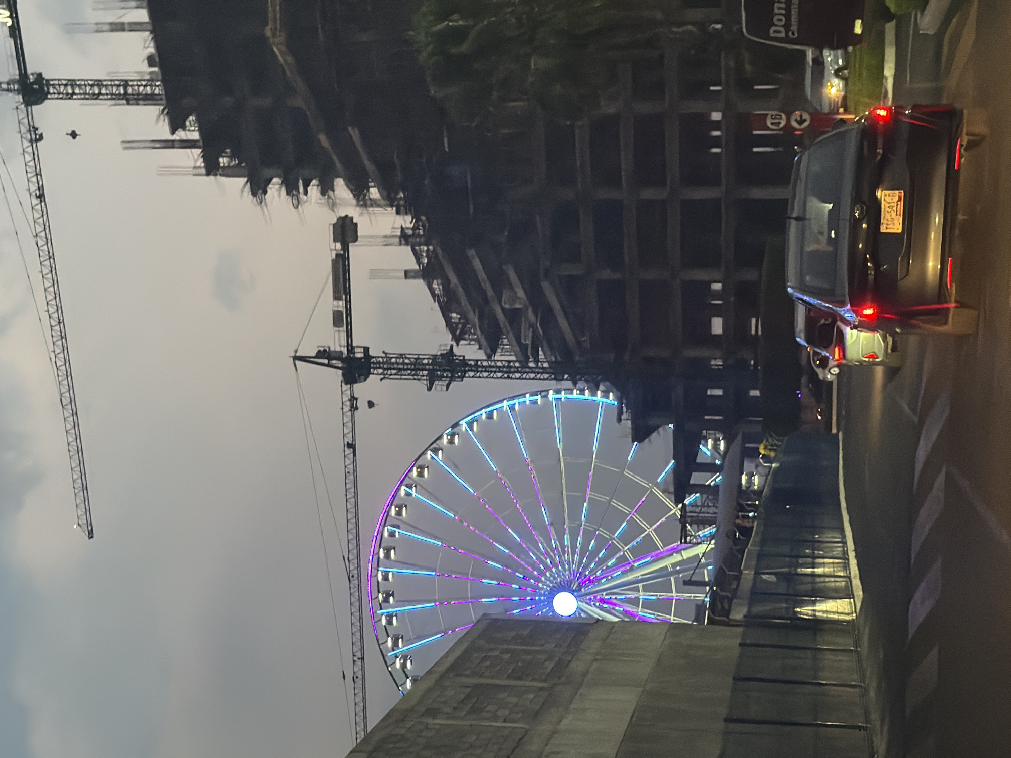 The image shows a scene in Puebla, Mexico, with a Ferris wheel illuminated with purple and blue lights as a focal point. The Ferris wheel is positioned between two construction cranes and near a building under construction. A dark sedan with its brake lights on is on the right side of the frame, along with part of another vehicle in front of it. The license plate of the sedan is visible and reads "T36-541-B". Buildings and trees can be seen in the background. The sky is overcast, indicating it is either early morning or late evening.
