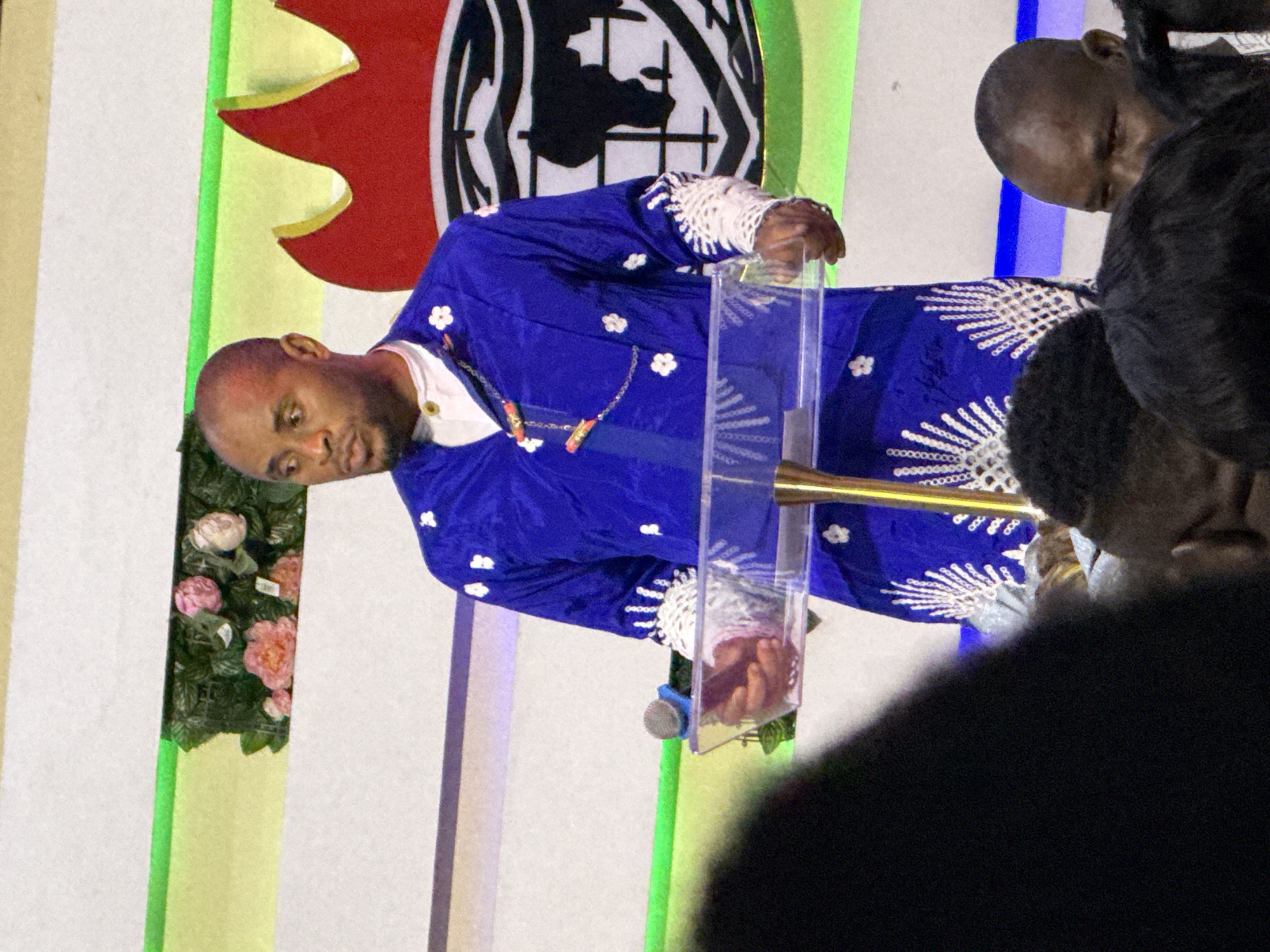 A man in a blue, flower-patterned robe stands behind a clear podium with a microphone, addressing a gathering in what appears to be a church setting. A group of individuals is situated to the right of the frame, partially obscured. Behind the speaker, there's a decorative backdrop featuring red and green elements, along with a black and white symbol. The location is Lugbe, Nigeria.