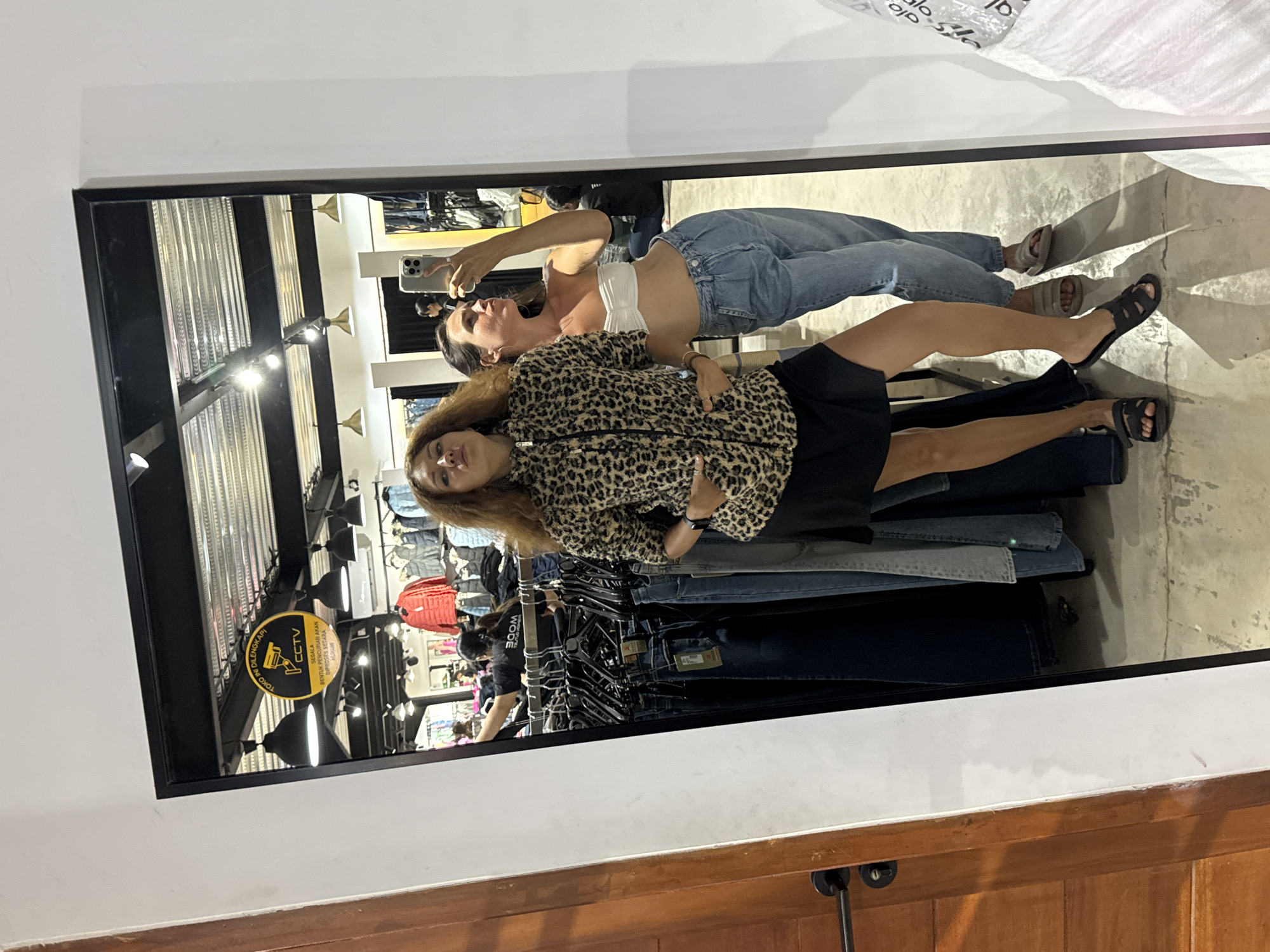 In the city of North Kuta, Indonesia, two women are posing for a mirror selfie in what appears to be a clothing store. The woman on the left is wearing a leopard-print jacket, a black skirt, and black sandals. She has her hands in her pockets and is looking at the camera with her lips pursed. The woman on the right is wearing a white strapless top, light-wash jeans, and grey sandals. She is holding a smartphone up to take the photo and is looking towards the mirror with her eyes closed and her mouth slightly open. Behind them, racks of clothing and a CCTV sign are visible. The floor is concrete and there are shadows cast by the people and clothing racks.