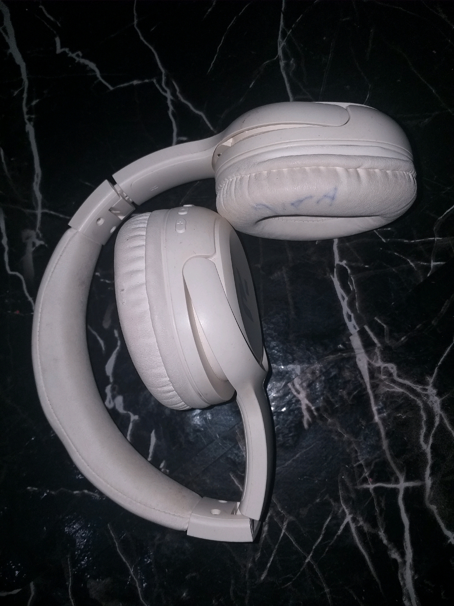 A pair of white, over-ear headphones is positioned on a dark, marble-patterned surface. The headphones are partially folded, with one earcup resting on the other. The surface appears to be a table or countertop. There are no people or other objects visible in the image. The lighting is dim, casting subtle shadows. The image is a close-up shot, focusing solely on the headphones and the immediate surface they are on. There is no indication of the location in Air Jamban, Indonesia, as the image contains no contextual clues related to a specific city or country.