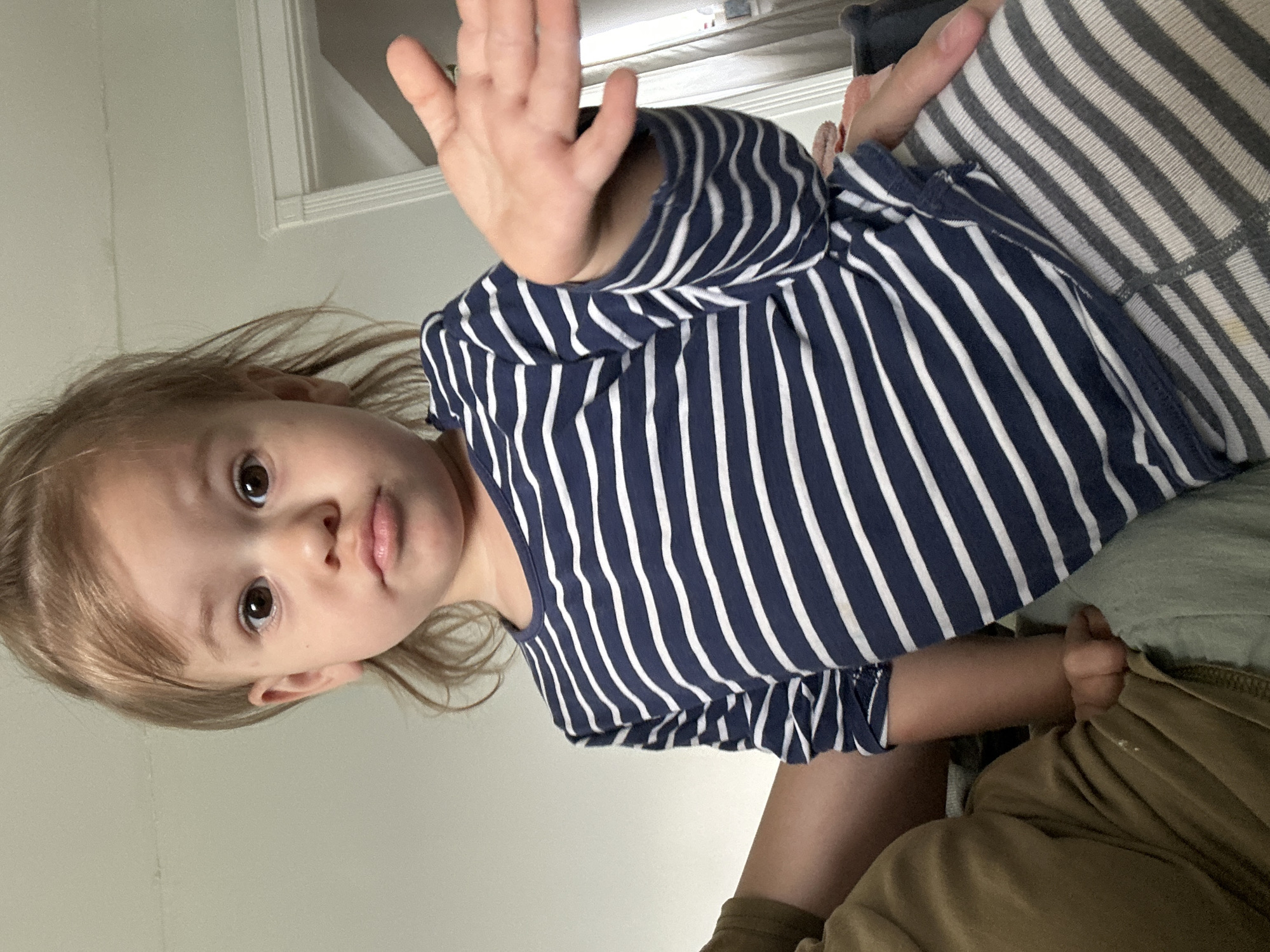 A young child is shown from a low angle indoors. The child has blonde hair and brown eyes, and is wearing a navy blue and white striped shirt. The child's right hand is raised with the palm facing the viewer. The child appears to be sitting or lying on a striped surface. Part of an adult's arm and khaki-colored clothing are also visible. The background appears to be a plain wall. The image does not contain any information that could identify the location in Kharnavy, Russia.