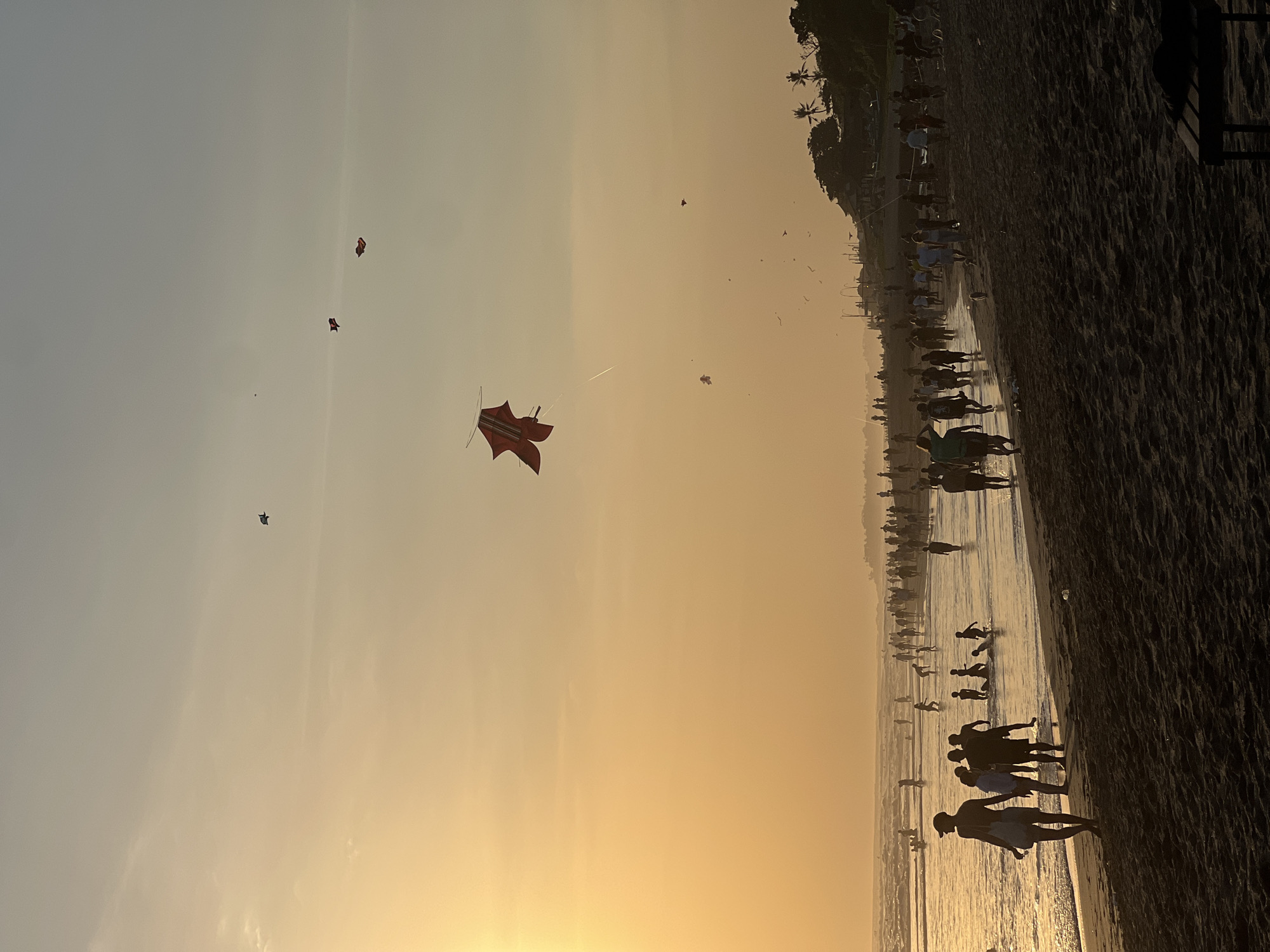 This image depicts a crowded beach scene in North Kuta, Indonesia, during sunset. The sky is a warm orange and yellow hue, with a few wispy clouds. Several kites are visible in the sky, including a large red and white kite in the foreground. Numerous people are walking along the shoreline, some wading in the shallow water, while others are enjoying the sand. The overall atmosphere appears to be lively and festive.