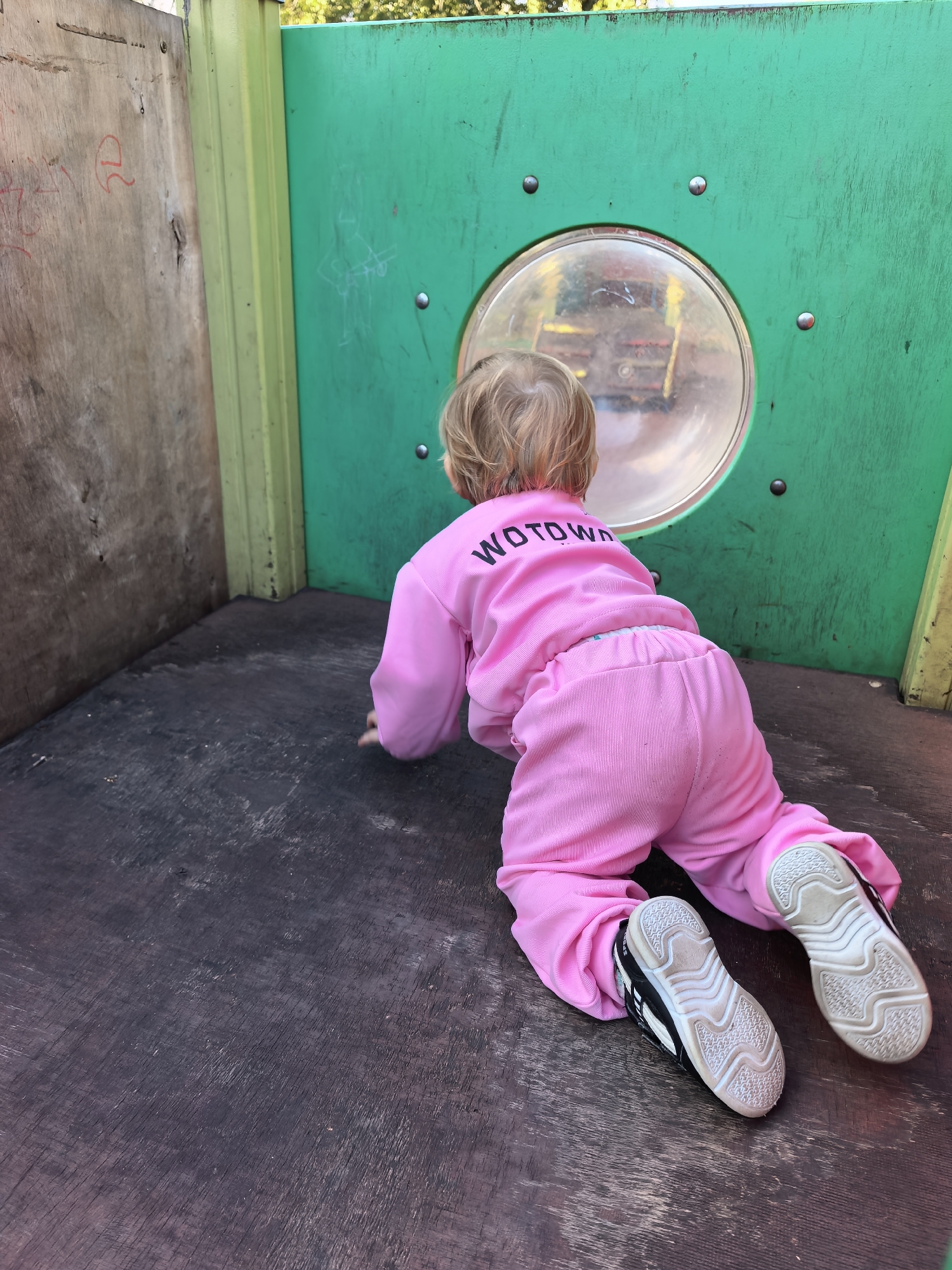 A young child wearing a pink tracksuit with the word "WOTOWN" printed in black letters on the back is crawling on a dark wooden floor inside a playground structure. The child is facing away from the camera and looking through a round, transparent window on a green metal panel. The window reflects an image of playground equipment. To the left of the green panel is a wooden panel with faint red markings. The environment appears to be an outdoor playground.