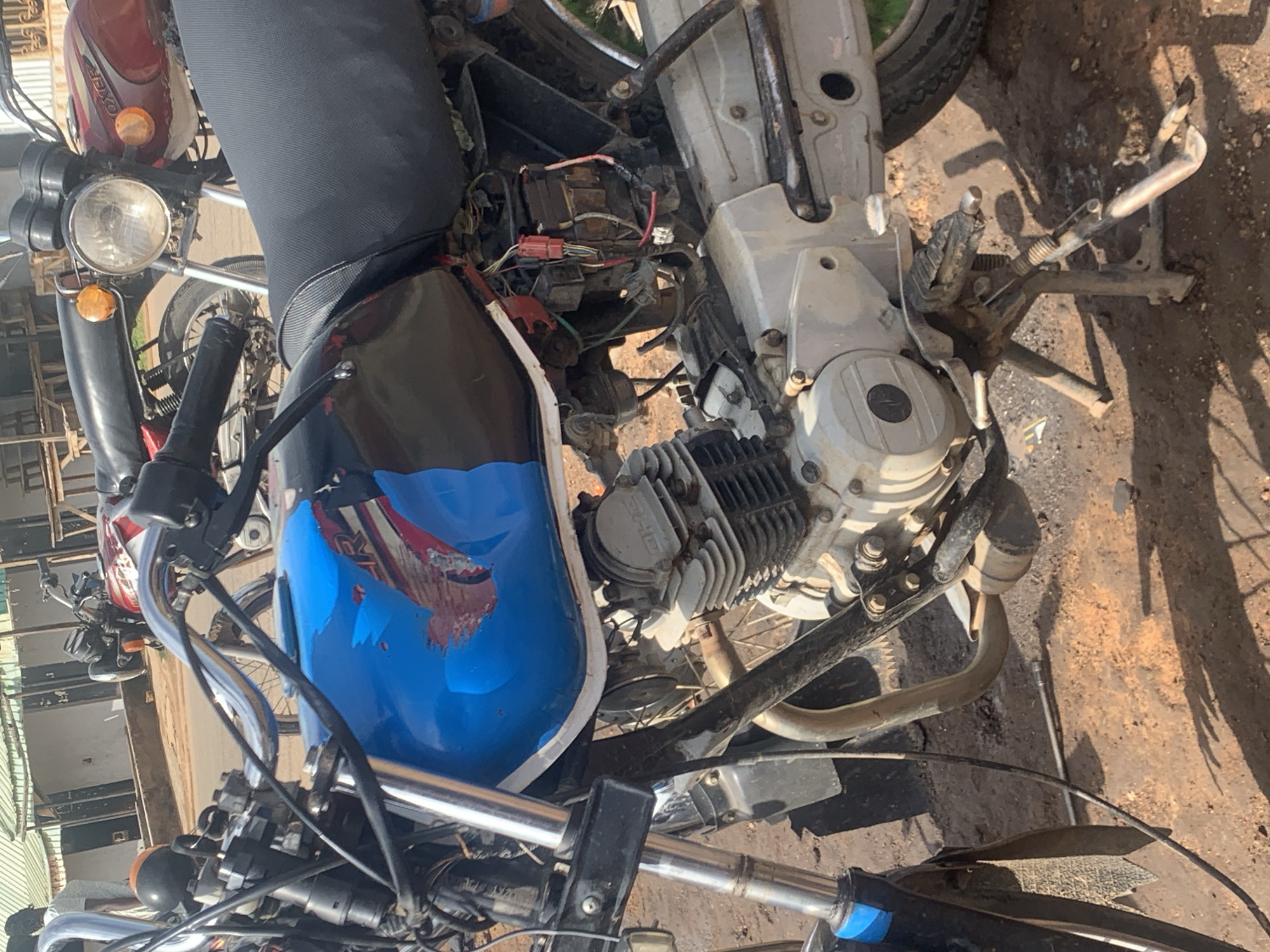 The image shows a close-up view of a blue and black motorcycle parked on a dirt ground in Gura Top, Nigeria. The motorcycle's engine, fuel tank, seat, handlebars, and wheels are visible. The fuel tank has a blue paint job with a graphic design. Wires and electrical components are exposed on the side of the motorcycle, suggesting it may be undergoing repairs or maintenance. Another motorcycle, red and black, is partially visible in the background. The environment appears to be an outdoor setting with a building and some wooden structures in the distance. The overall scene depicts a common sight in the region, where motorcycles are a prevalent mode of transportation.