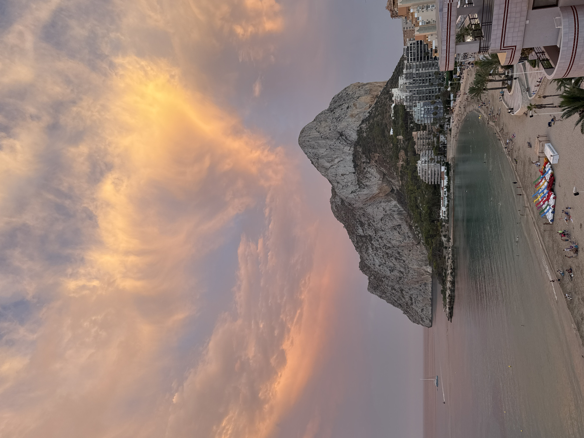 The image depicts a scenic coastal view in Calp, Spain, featuring the iconic Penyal d'Ifac rock formation. The sky above is filled with dramatic, colorful clouds, illuminated by the soft light of either sunrise or sunset, casting hues of orange, pink, and yellow across the scene. Below the sky, the Penyal d'Ifac dominates the mid-ground, a massive, rocky prominence jutting out into the Mediterranean Sea. The lower slopes of the rock are covered in greenery, and the base is dotted with buildings, suggesting a town nestled against the natural landmark.
The foreground shows a sandy beach with several people scattered across it, some walking, some sitting, and some swimming in the calm, greenish waters of the bay. Numerous colorful paddle boats are lined up on the sand, ready for use. Along the beach, a promenade is visible, lined with palm trees and multi-story buildings, indicative of a resort town. A white sailboat with its sails down is seen in the distance on the water to the left of the image. The overall impression is one of a peaceful, picturesque coastal setting, likely captured during a pleasant evening or morning.