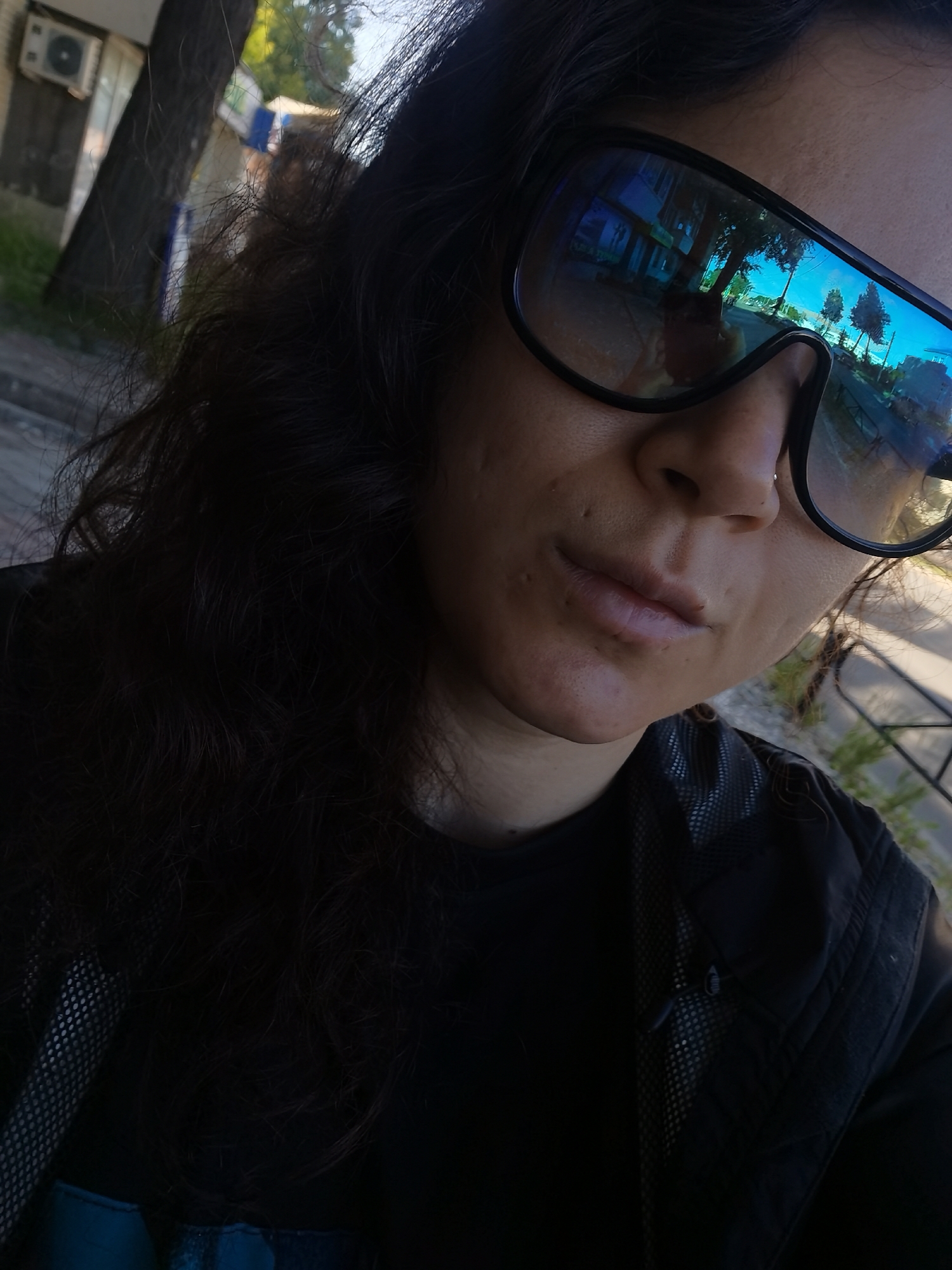 The image is a close-up, selfie-style photograph of a person wearing large, dark sunglasses with a blue tint. The person has dark, wavy hair and is wearing a black t-shirt and a black jacket. The background is out of focus but appears to be an outdoor urban setting with buildings, trees, and a street visible in the reflection of the sunglasses. There is an air conditioning unit mounted on a building in the upper left corner of the image. The overall lighting suggests it is daytime. The specific location in the city of Chaikovsky, Russia, cannot be determined from the visual information provided.