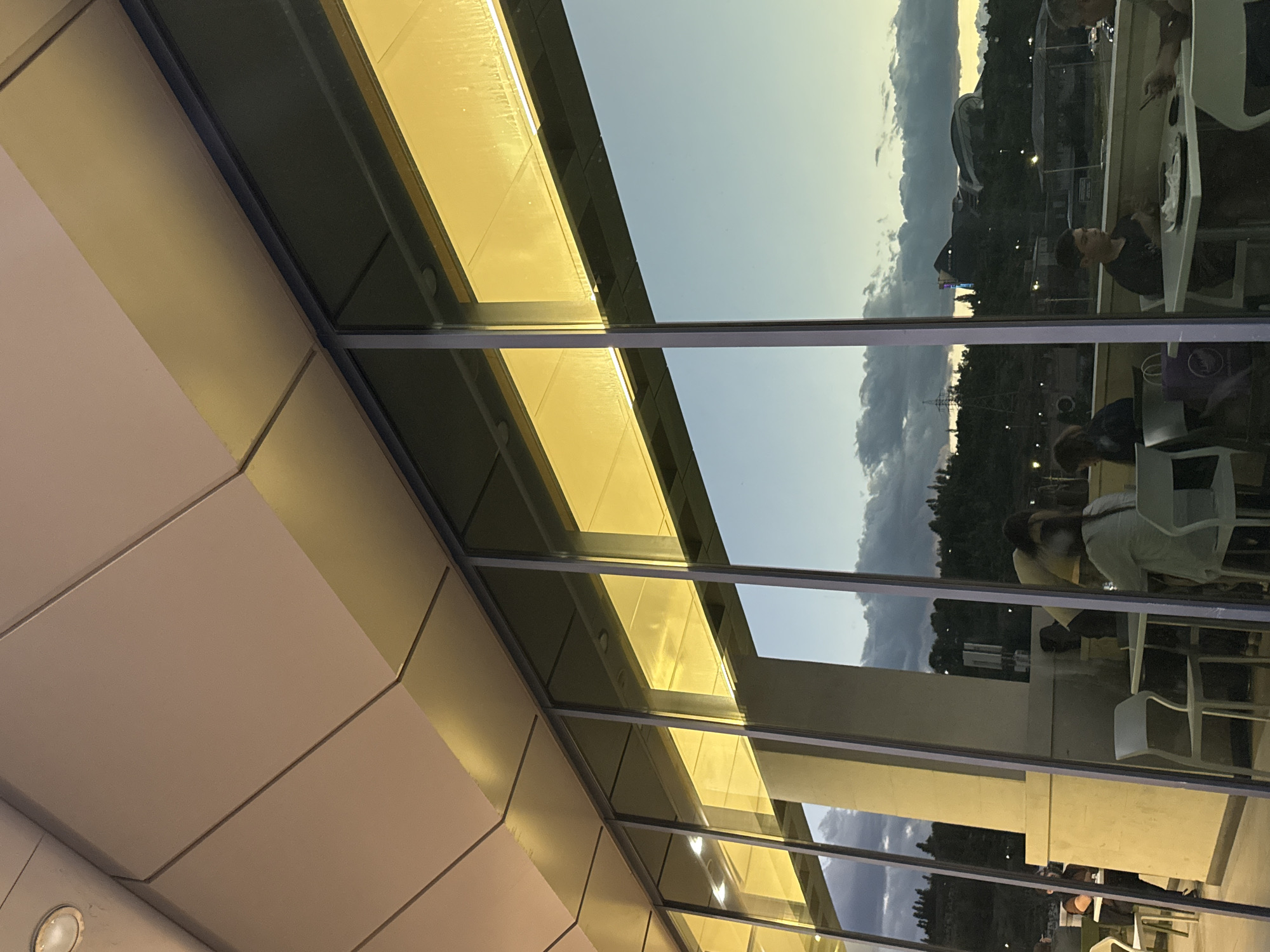 The image, when viewed in its correct orientation, depicts a modern architectural scene featuring a building facade with large glass panels. On the left, the building is composed of light-colored, possibly beige or off-white, panels arranged with distinct horizontal and diagonal lines. Above and reflected in the glass, a section of the building glows with a warm, golden-yellow light, likely from internal illumination or a reflective surface catching the sun.
The extensive glass panels act as both windows and reflective surfaces, showcasing an outdoor environment. Through the glass and in its reflections, a soft blue sky with scattered grey-white clouds is visible, suggesting either late afternoon/early evening or early morning. In the distant background, a hilly landscape is silhouetted, featuring trees, various urban structures, and a prominent large building or complex on a hill. Utility poles or towers are also seen in the middle ground.
In the reflected foreground, several individuals are seated at outdoor tables and chairs, indicating a cafe or restaurant setting. One person in a dark t-shirt is clearly visible sitting at a white table, while other figures, including a person's head and shoulders, are seen at adjacent tables. The activity appears to be casual dining or socializing. There is no visible text in the image. The scene captures a moment of leisure at a transitional time of day in Yerevan, Armenia.