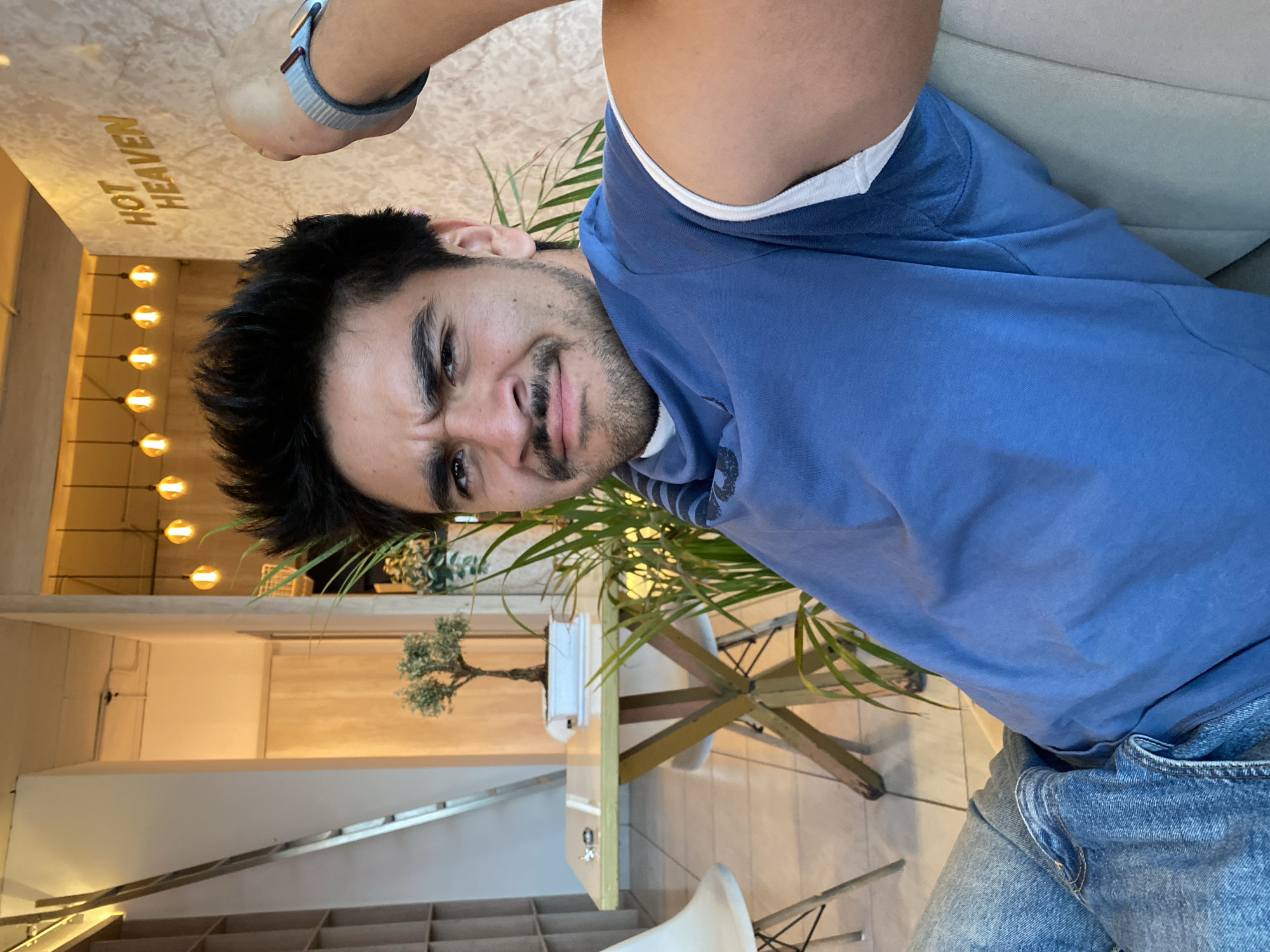 A young man is featured in an indoor portrait, seated in what appears to be a cafe or contemporary establishment in Puebla, Mexico. He looks directly at the camera with a playful or slightly quizzical expression, characterized by furrowed brows and a faint smirk. He has dark hair and a beard, and is dressed in a blue long-sleeved top over a white undershirt, paired with blue denim jeans. A light blue and white striped watch band is visible on his right wrist.
The setting includes warm, ambient lighting from several spherical fixtures mounted vertically on a wall in the background. A table, topped with a small, bonsai-like plant, is situated near a larger, leafy green plant that partially screens the distant background. A staircase or shelving unit is visible on the left side of the frame. The words "HOT HEAVEN" are prominently displayed in gold lettering on a light, textured surface above, suggesting the name of the venue. The overall atmosphere appears modern and inviting.