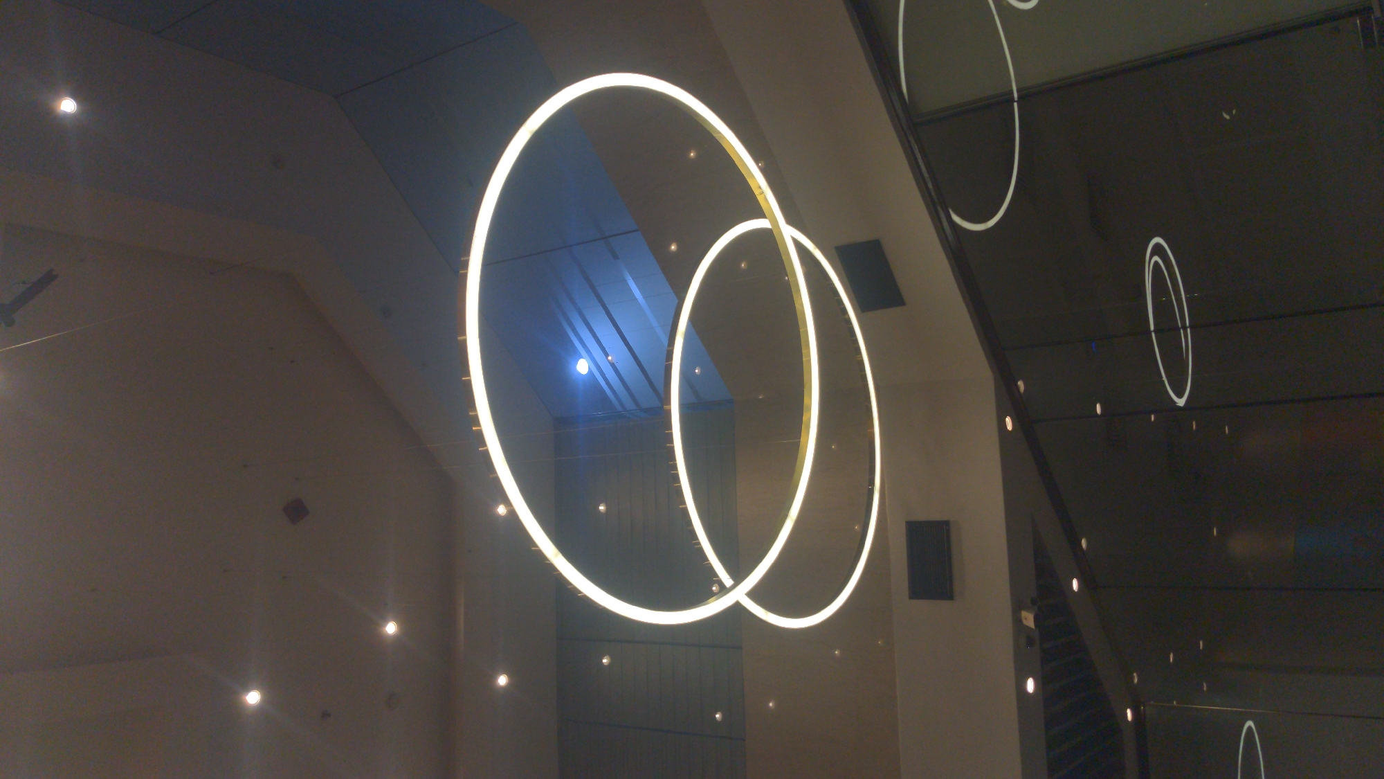 This image captures a modern interior space, characterized by prominent circular lighting fixtures. Two large, intertwined or concentric rings, emitting a bright, warm white light, are suspended as central decorative elements. Smaller, circular spotlights are visible on the light-colored ceiling and walls, contributing to the overall illumination.
A significant feature is the dark, highly reflective surface on the right side of the frame, which strongly mirrors the primary light rings and additional spotlights. These reflections appear as ghostly white circles and dots, extending the lighting pattern across the surface. Some areas of the ceiling also exhibit a bluish tint, possibly from accent lighting or ambient reflections.
The scene is devoid of people or specific activity, focusing instead on the architectural and lighting design. The ambiance is contemporary and sophisticated, suggesting a modern building or an artistic installation. The time of day is implied to be evening or night due to the prominence of artificial lights. No text is visible in the image. The location is contextualized as Abuja, Nigeria.
