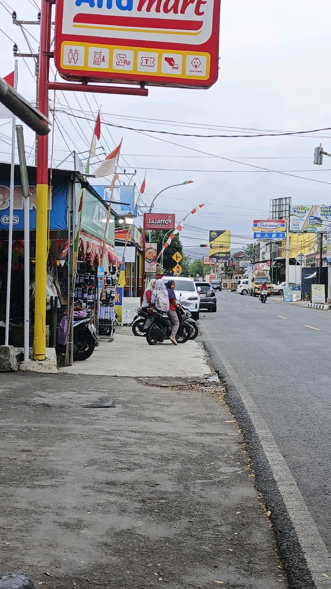 The image captures a street scene in Desa Karangbenda, Indonesia, characterized by a roadside with shops and a busy road. On the left, a prominent red and white sign for "Alfamart" is visible, adorned with various icons. Adjacent to it are small shops with awnings and displays of goods, including what appears to be phone accessories. Two individuals wearing hijabs are seated on a dark-colored motorcycle parked on the sidewalk in front of a shop. Another motorcycle is parked behind them. The road itself is asphalt, with a white dividing line and appears to have moderate traffic, including cars and motorcycles. In the background, a range of businesses and billboards line the street, with mountains visible in the distance under an overcast sky. The overall impression is of a vibrant commercial area within a semi-urban or rural Indonesian setting.
