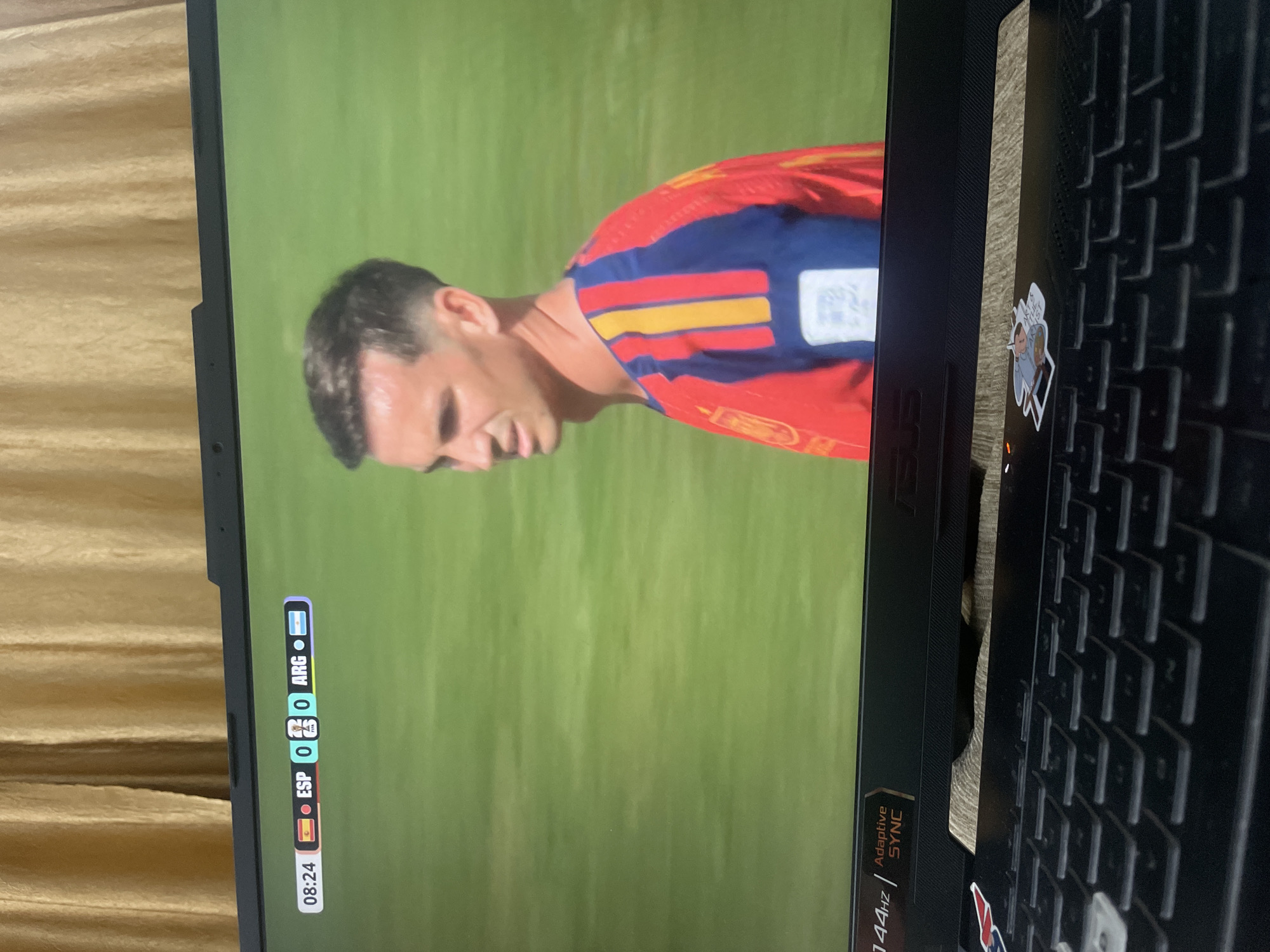 A young man in a red and blue striped soccer jersey is on a bright green field. He is looking to his right, with his mouth slightly open. The score on the screen reads "08:24 ESP 0 0 ARG." The screen is part of a laptop, with a portion of the keyboard visible in the foreground. The background of the image is a tan curtain. The scene is indoors, and it is unlikely to be in Bhagsu, India, as the score display suggests a sporting event, not a typical Bhagsu location.