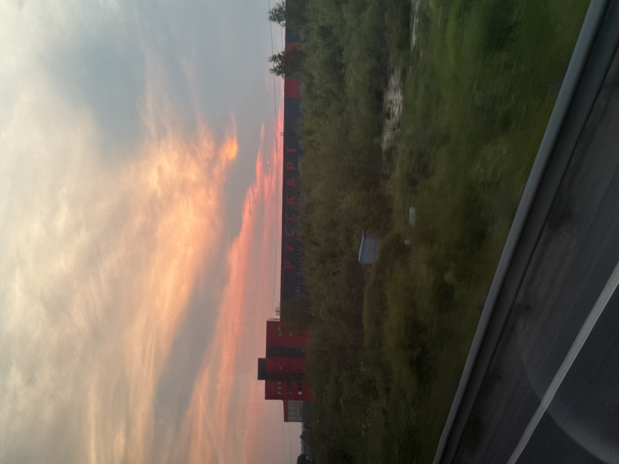 The image depicts a large industrial building in Nikol'skoe, Russia, silhouetted against a sunset sky. The building, painted in a striking combination of red and dark gray, has a prominent red section with multiple windows and a darker section with large lettering that appears to read "РУССКАРТ". A dense line of green trees and bushes separates the foreground from the building. In the foreground, a dark gray barrier with a white stripe, likely a highway guardrail, suggests the perspective is from a moving vehicle. The sky is filled with dramatic clouds, painted in hues of orange, pink, and gray, indicative of twilight. A small blue structure is visible amidst the greenery. There are no people visible in the image.