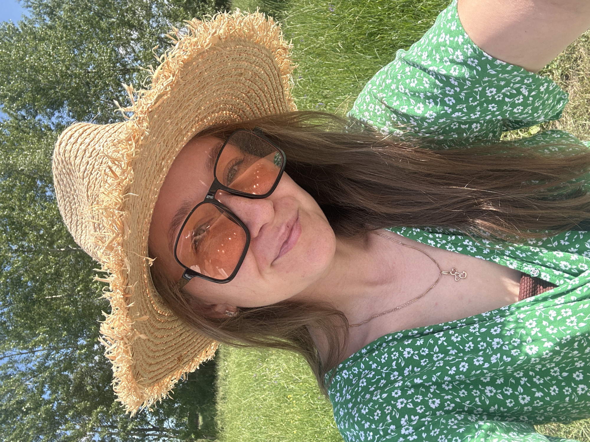 A woman is sitting in a grassy field, possibly in Bely Yar, Russia, as suggested by the metadata. She is wearing a straw hat with frayed edges, large square-framed orange-tinted sunglasses, and a green floral-patterned dress. A thin gold necklace with a small, stylized cross pendant is visible around her neck. Her hair is long and brown. The background consists of green trees and a bright blue sky. She is looking at the camera and smiling slightly. The lighting indicates it is a sunny day.